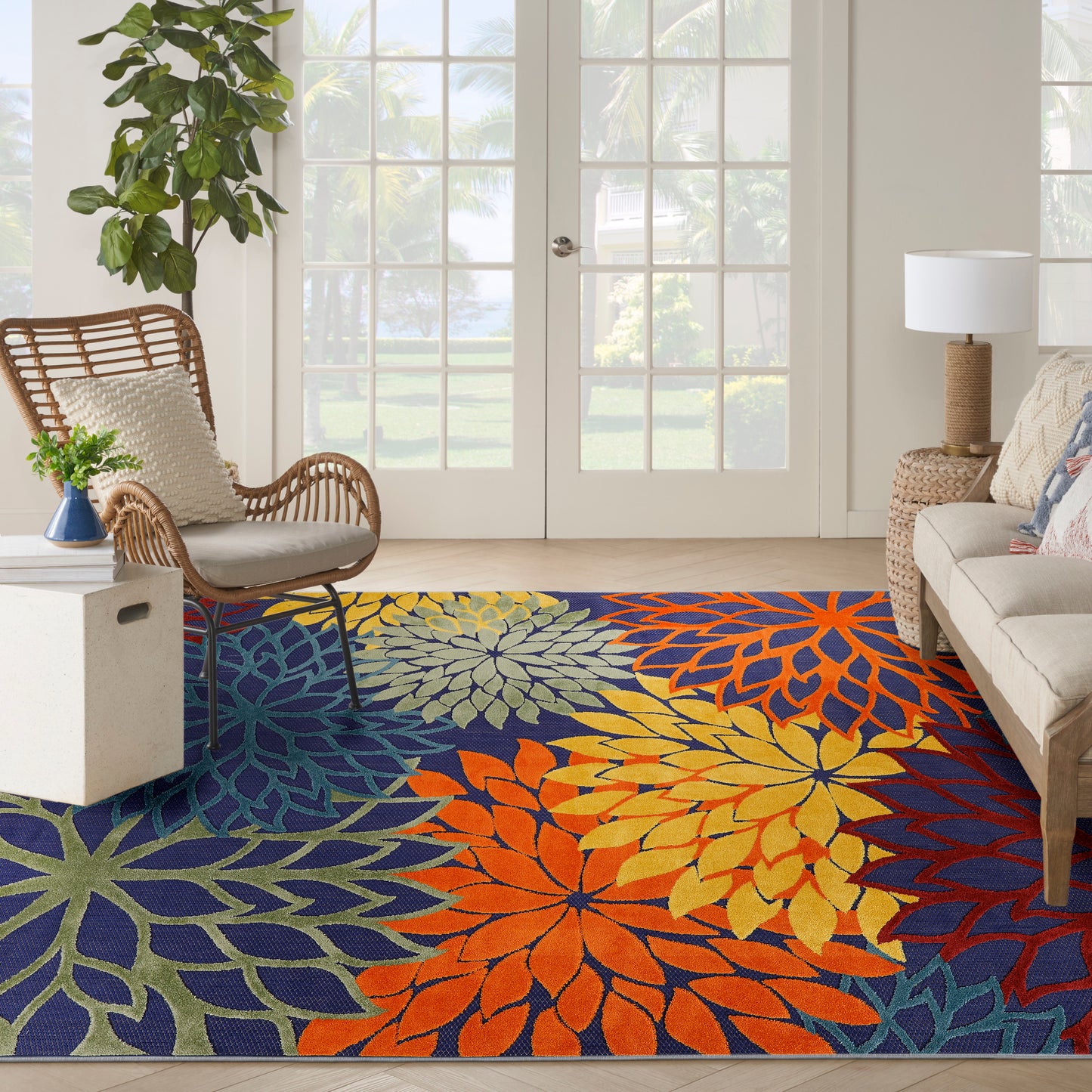 Vibrant multicolor outdoor floral rug in blue, orange, yellow and green; wicker chair, beige sofa, leafy plant, glass doors.