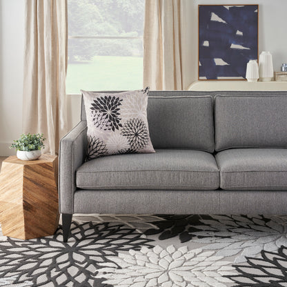Gray fabric sofa with a floral pillow, geometric wooden side table, beige curtains, blue abstract art, and a floral rug.
