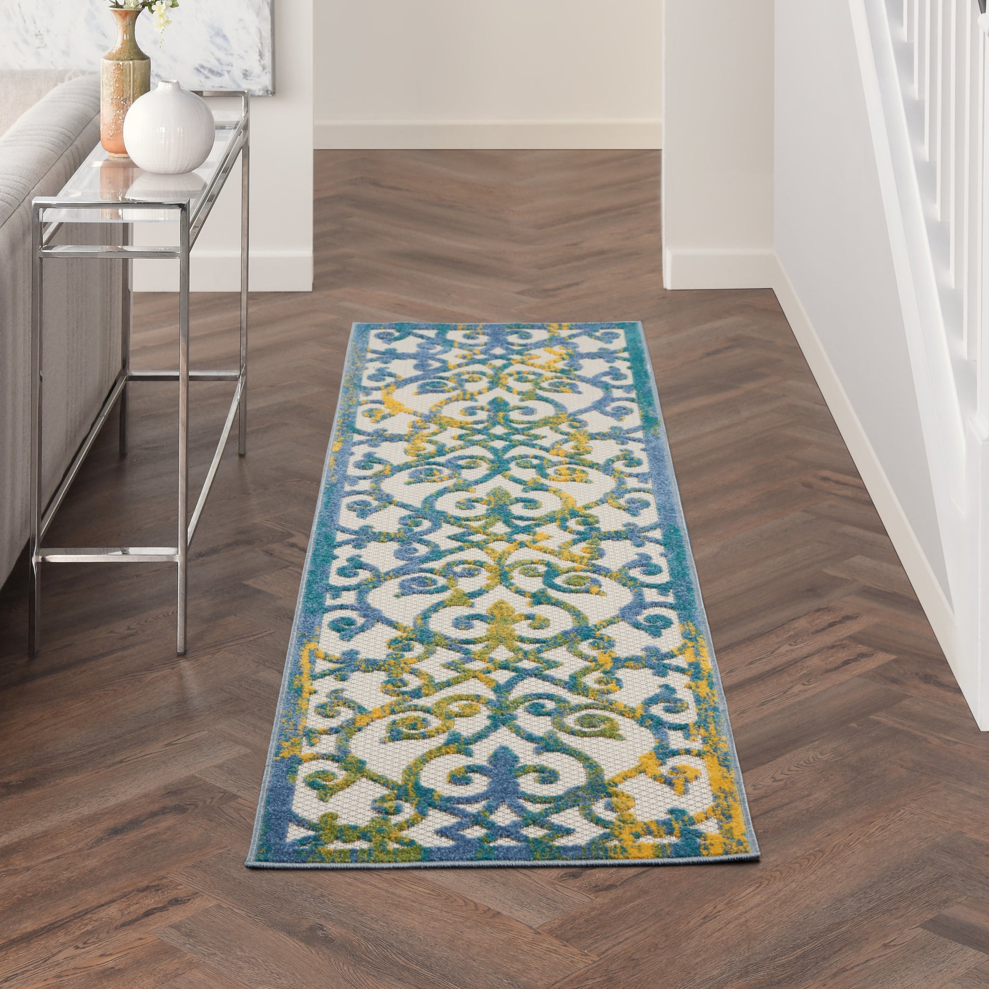 Long rectangular rug runner with blue, teal, and yellow scrollwork in a modern hallway beside a glass-console table.