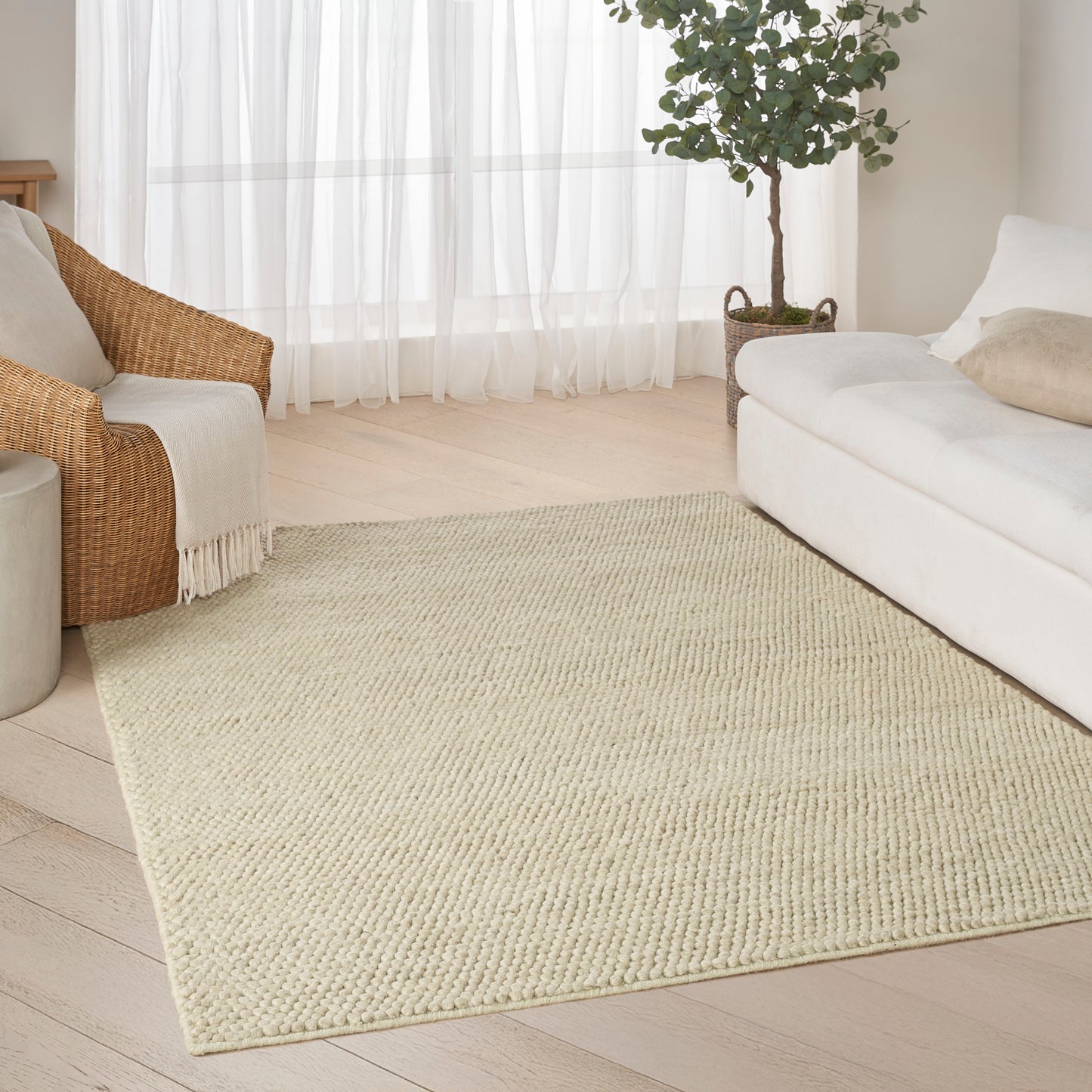 Neutral beige woven jute rug in a bright living room with a white sofa, wicker chair, sheer curtains, and a potted plant