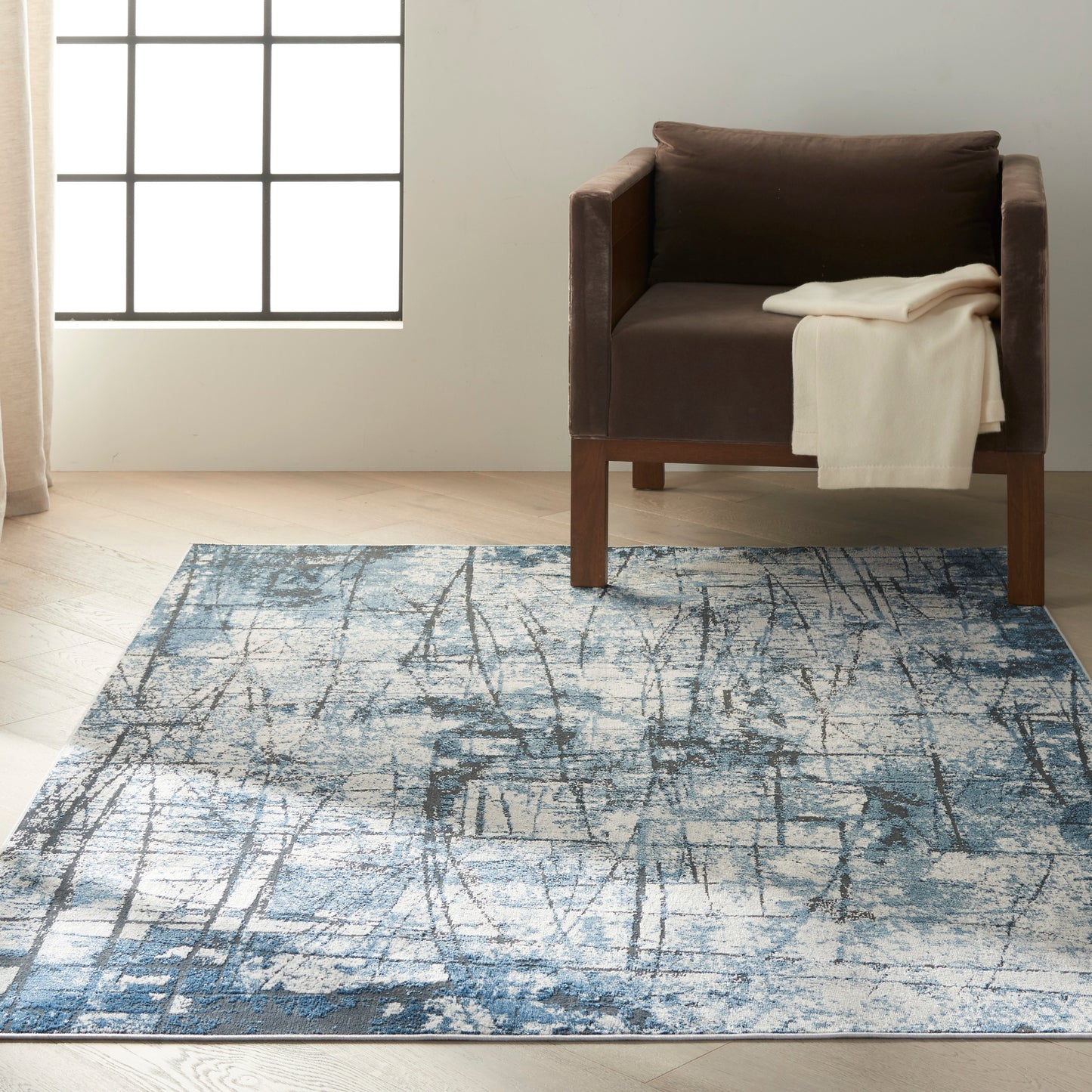 Brown upholstered armchair with wooden legs, cream throw, on a blue abstract-pattern rug in a sunlit room.