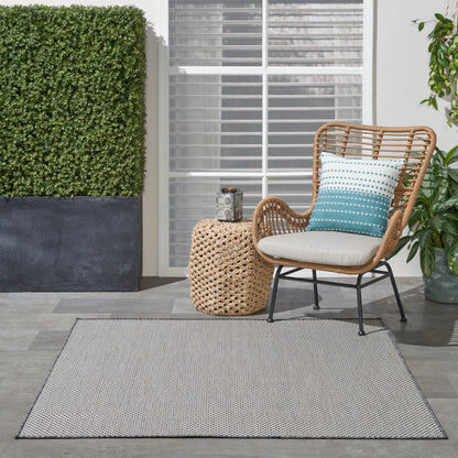 Outdoor wicker chair with white cushion and teal striped pillow, round woven side table, potted plants, and gray outdoor rug.
