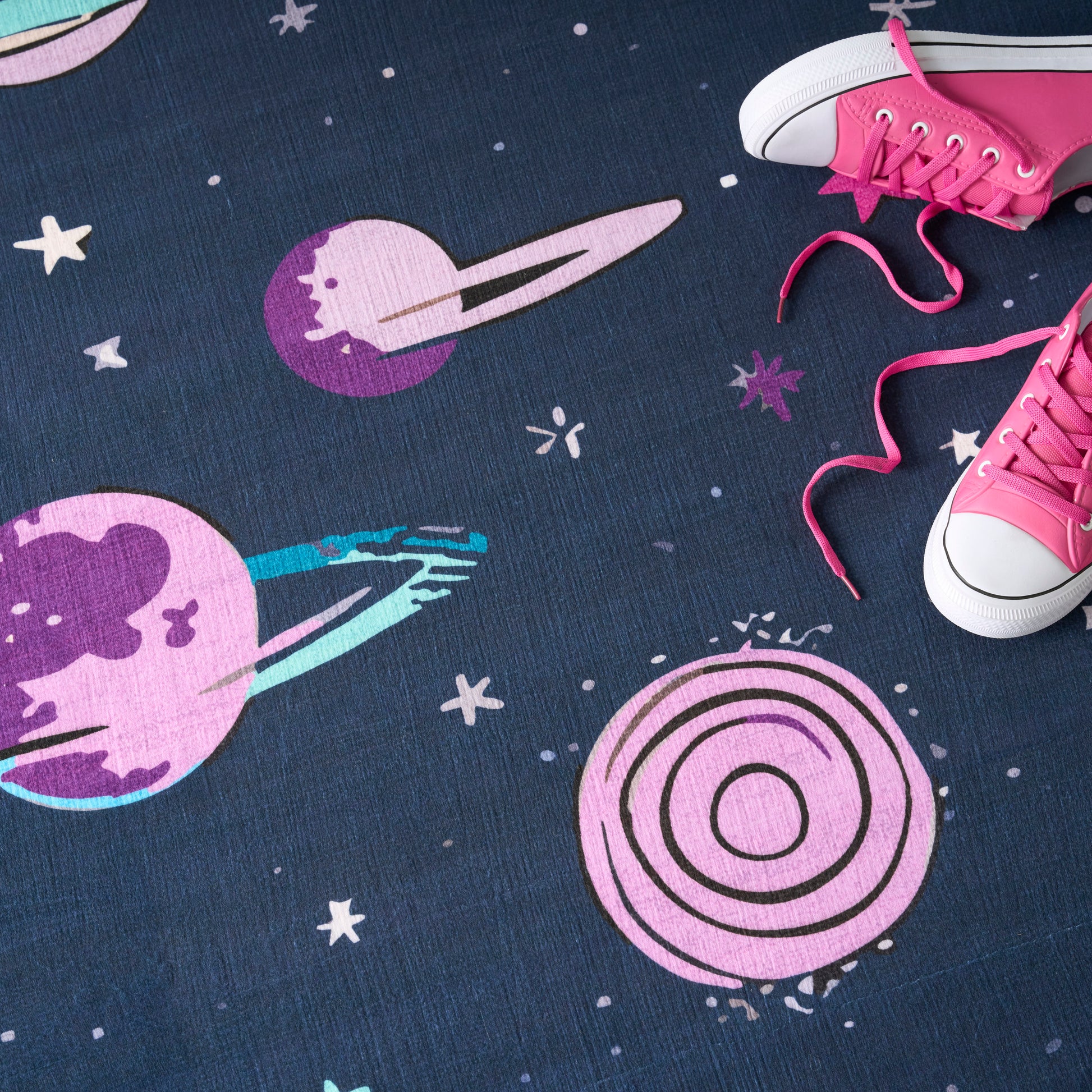 Space-themed blue rug featuring pink and purple planets, rings and stars; pink sneakers with white toe caps sit at the edge.