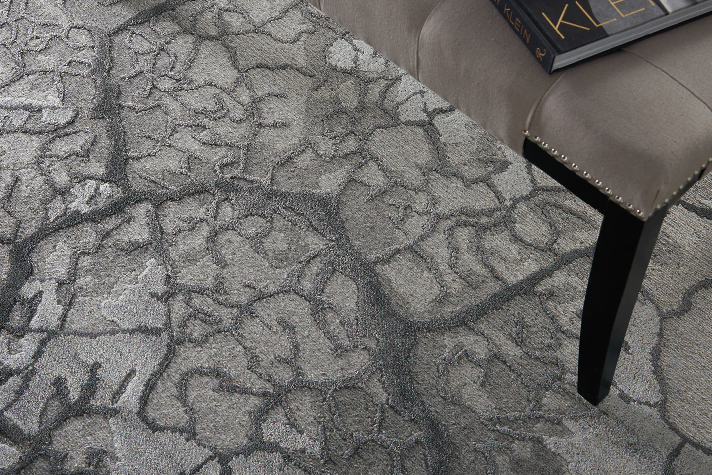 Gray textured area rug with an abstract pattern; beige tufted stool with studded trim and black tapered leg, with a book on top.