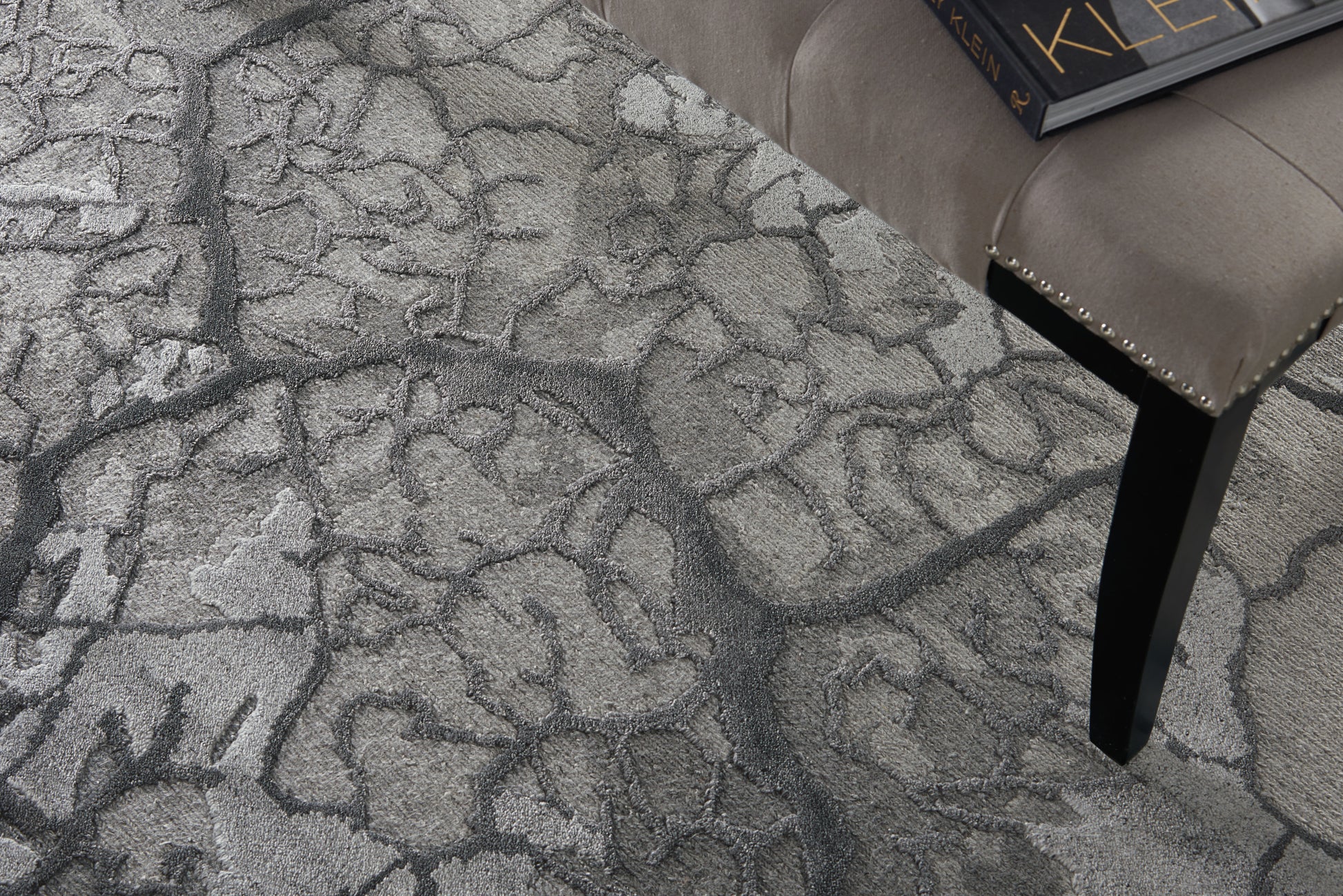 Gray textured area rug with an abstract pattern; beige tufted stool with studded trim and black tapered leg, with a book on top.