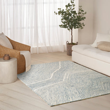 Light living room with pale blue chevron rug on light wood floor, white sofa, wicker chair, potted plant, sheer curtains.
