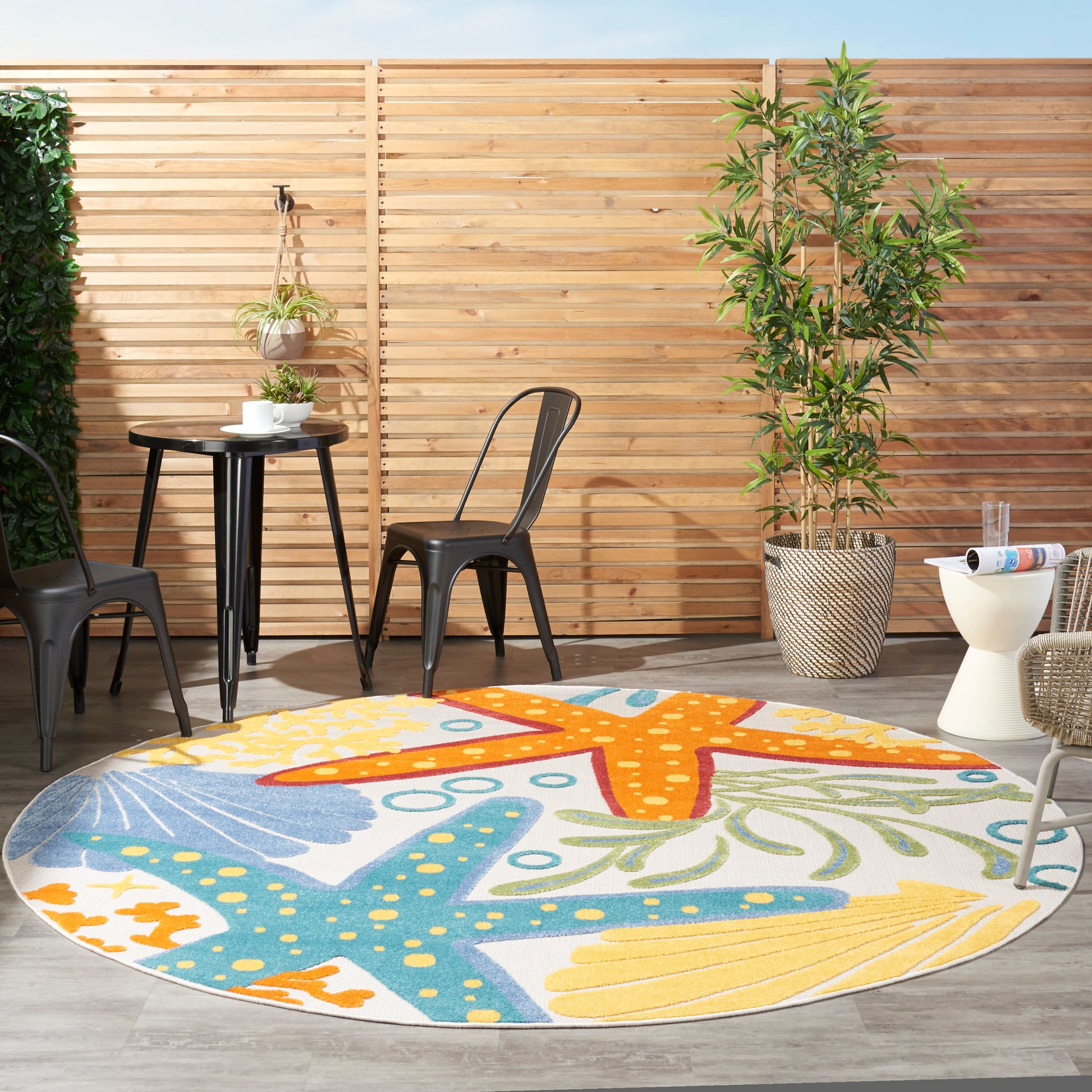 Round outdoor rug with orange, blue and yellow starfish patterns on a wooden deck, with black metal chairs and potted plants.