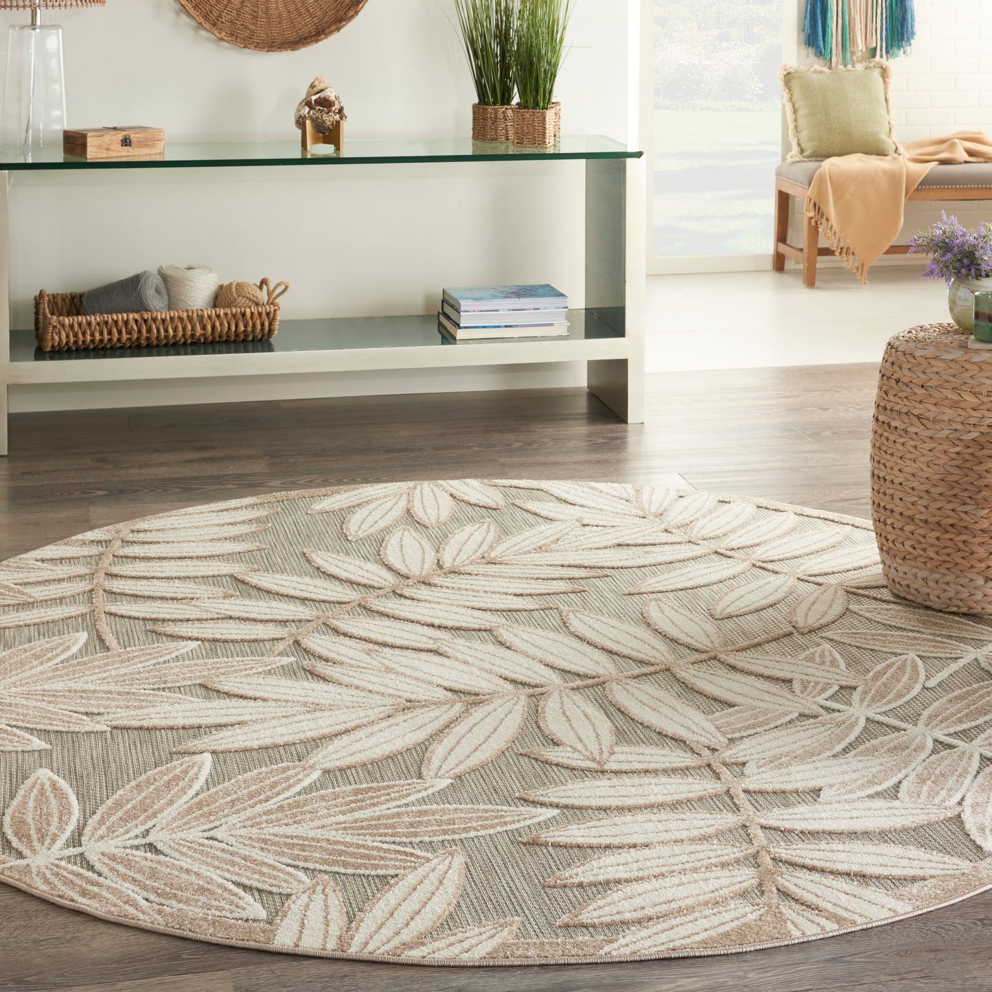 Round beige woven rug with white leaf pattern in a bright boho living room with a glass console, wicker basket, and plants.