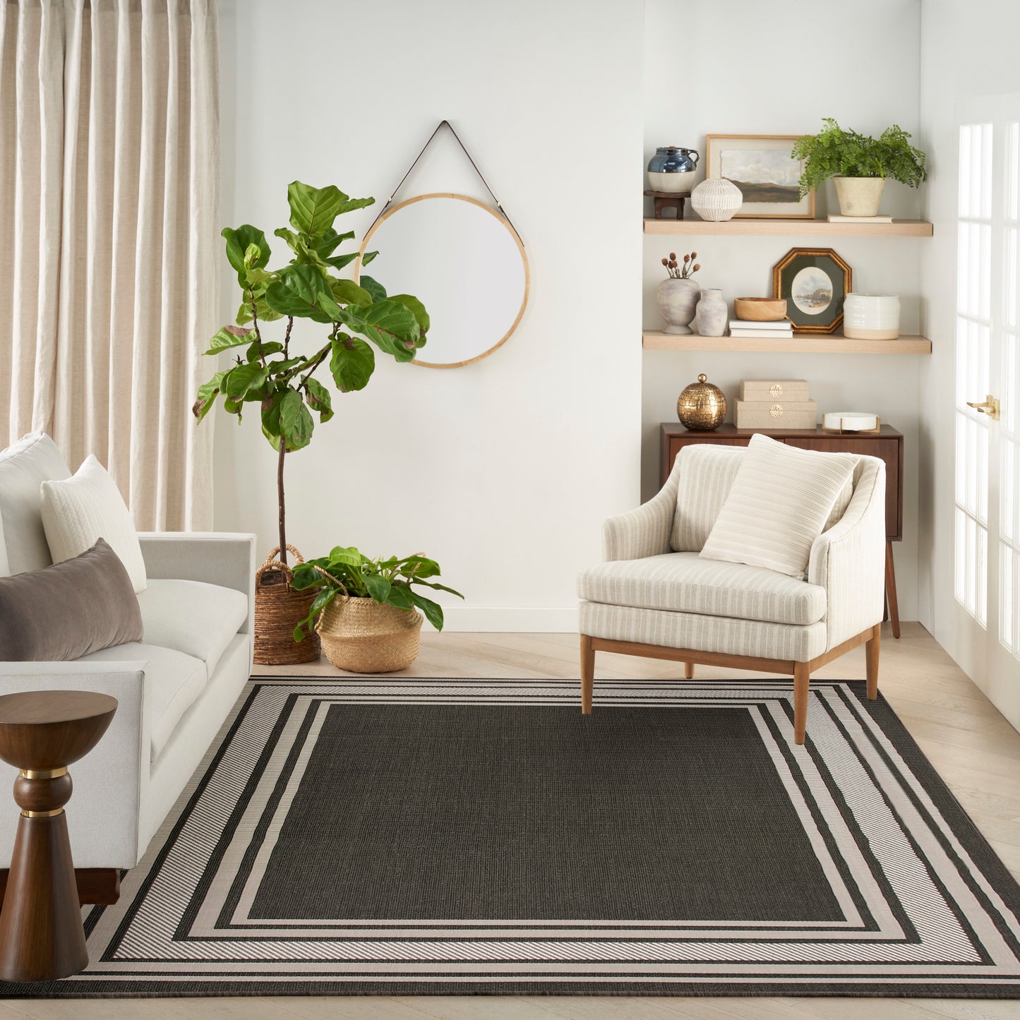 Neutral living room with beige sofa, striped armchair, large plant, round wall hoop, wood shelves & layered gray area rug.