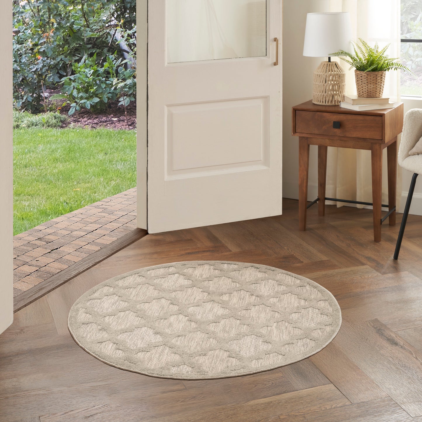 Round beige tufted rug on wooden floor in an entryway, open white door, with wood side table, lamp and plant nearby.