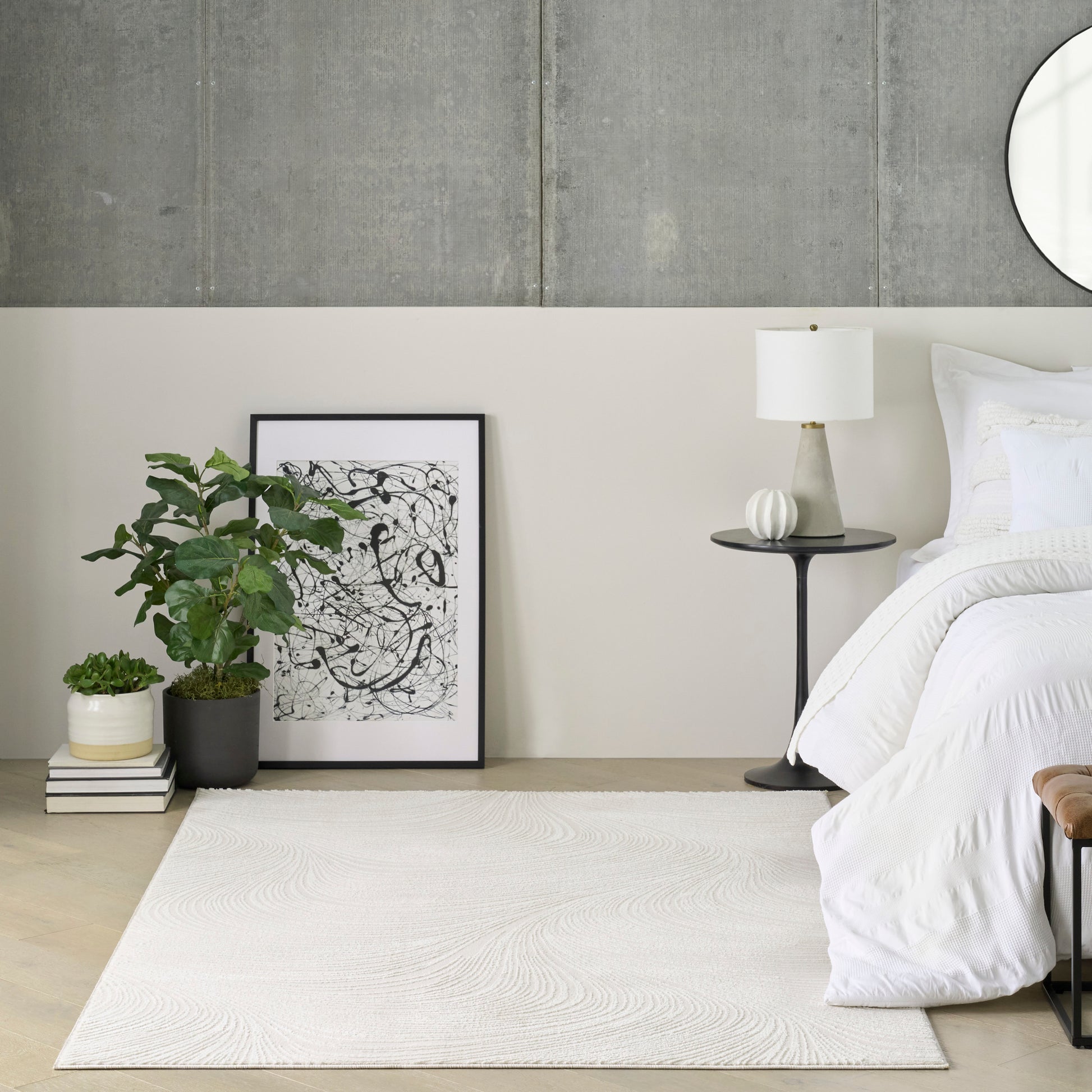 Minimalist bedroom with white bedding, concrete wall, mirror, abstract art leaning on floor, plant, lamp, cream rug.