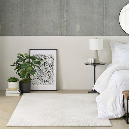 Minimalist bedroom with white bedding, concrete wall, mirror, abstract art leaning on floor, plant, lamp, cream rug.