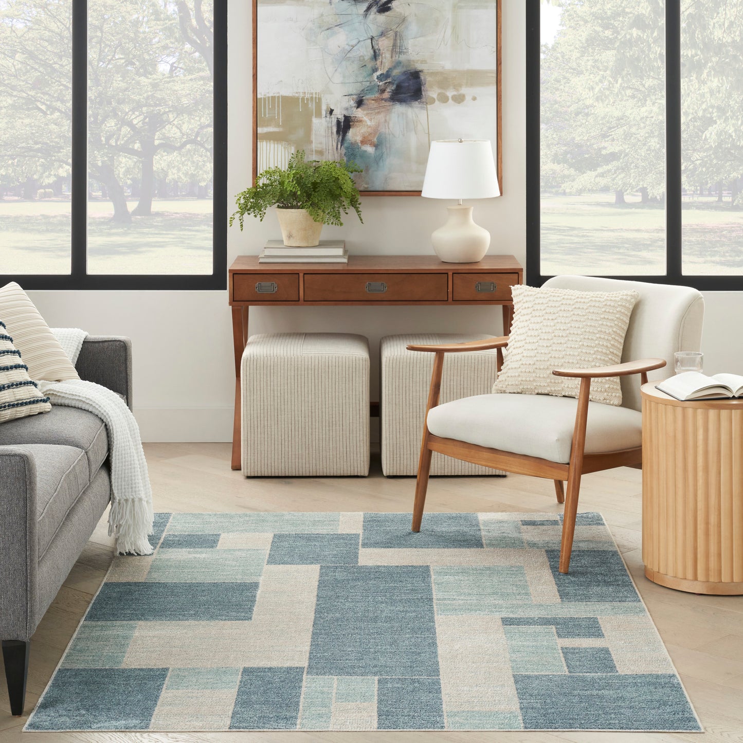 Blue geometric patchwork rug anchors a bright living room with a mid-century chair, wood console, ottomans, lamp.