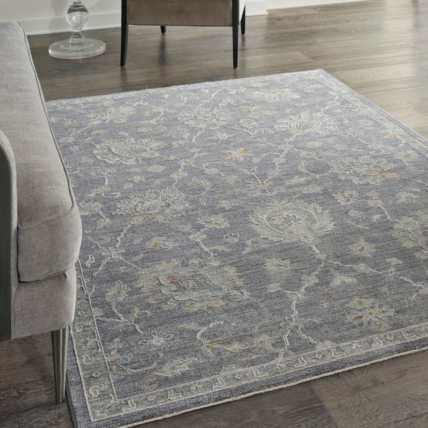 Blue-gray ornate area rug with faded floral pattern and light border on dark wood floor beside a gray sofa