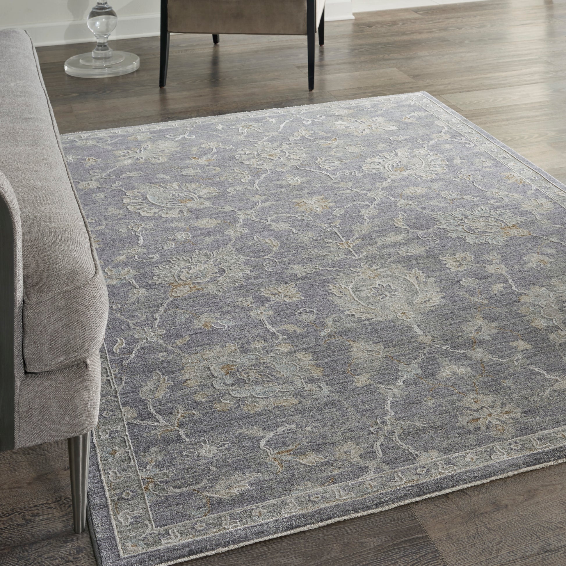 Blue-gray ornate area rug with faded floral pattern and light border on dark wood floor beside a gray sofa