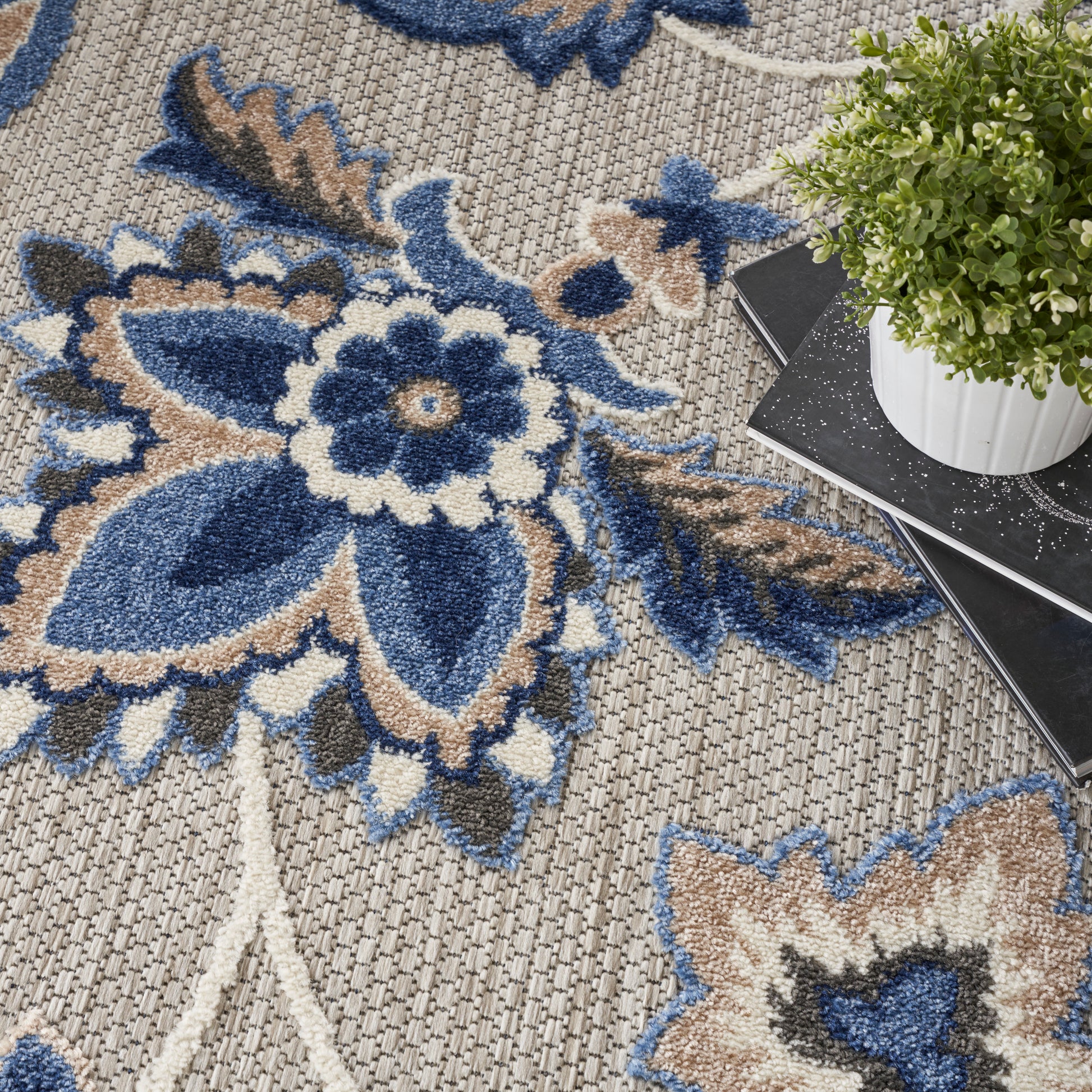 Blue, beige, and cream floral area rug with woven texture; large blue flower motif and potted plant on dark notebook.