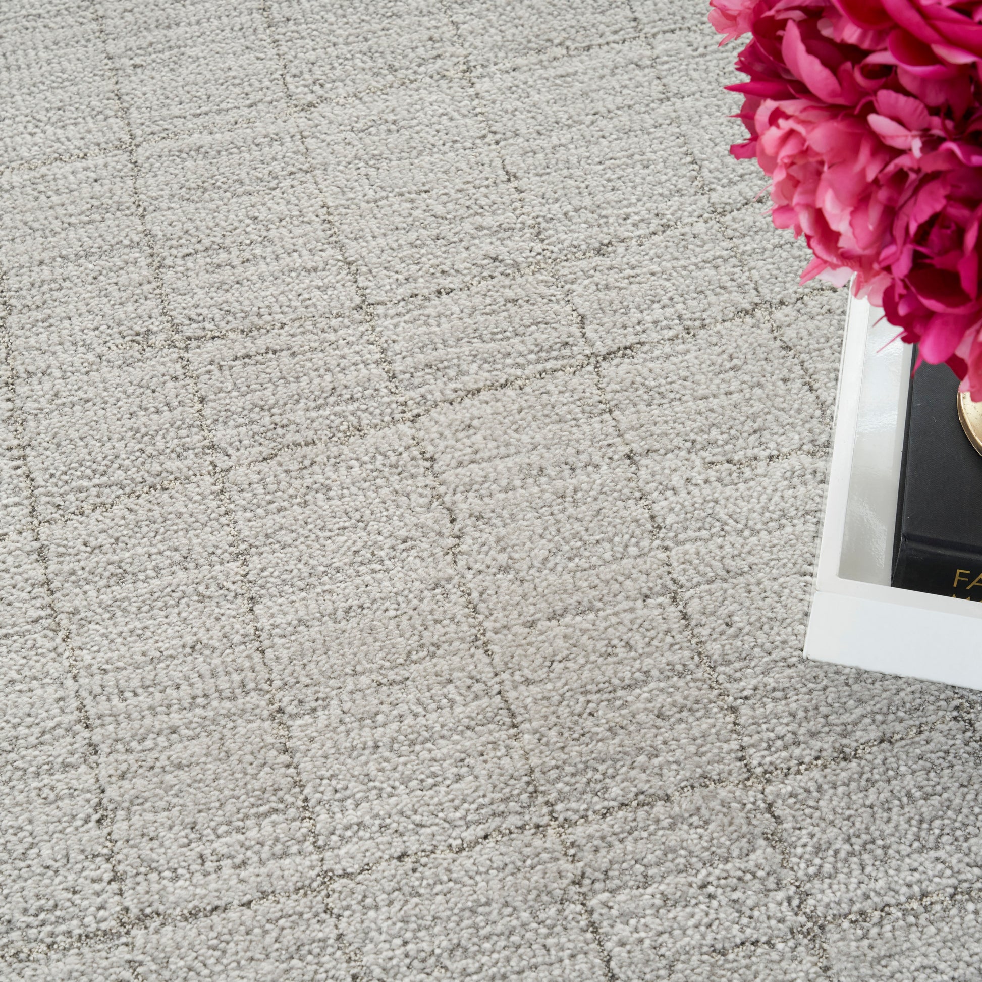 Beige textured rug with a subtle grid pattern; bright pink peonies in a white tray beside a black book.