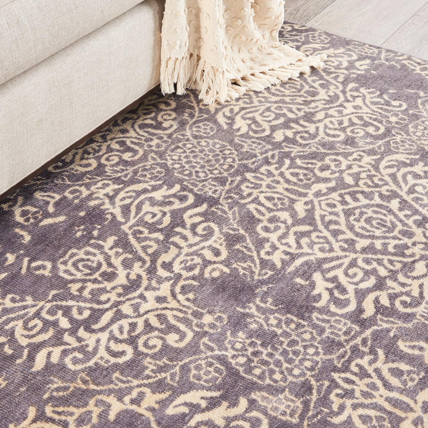 Purple and cream damask pattern rug beside a beige sofa with a cream fringed throw.