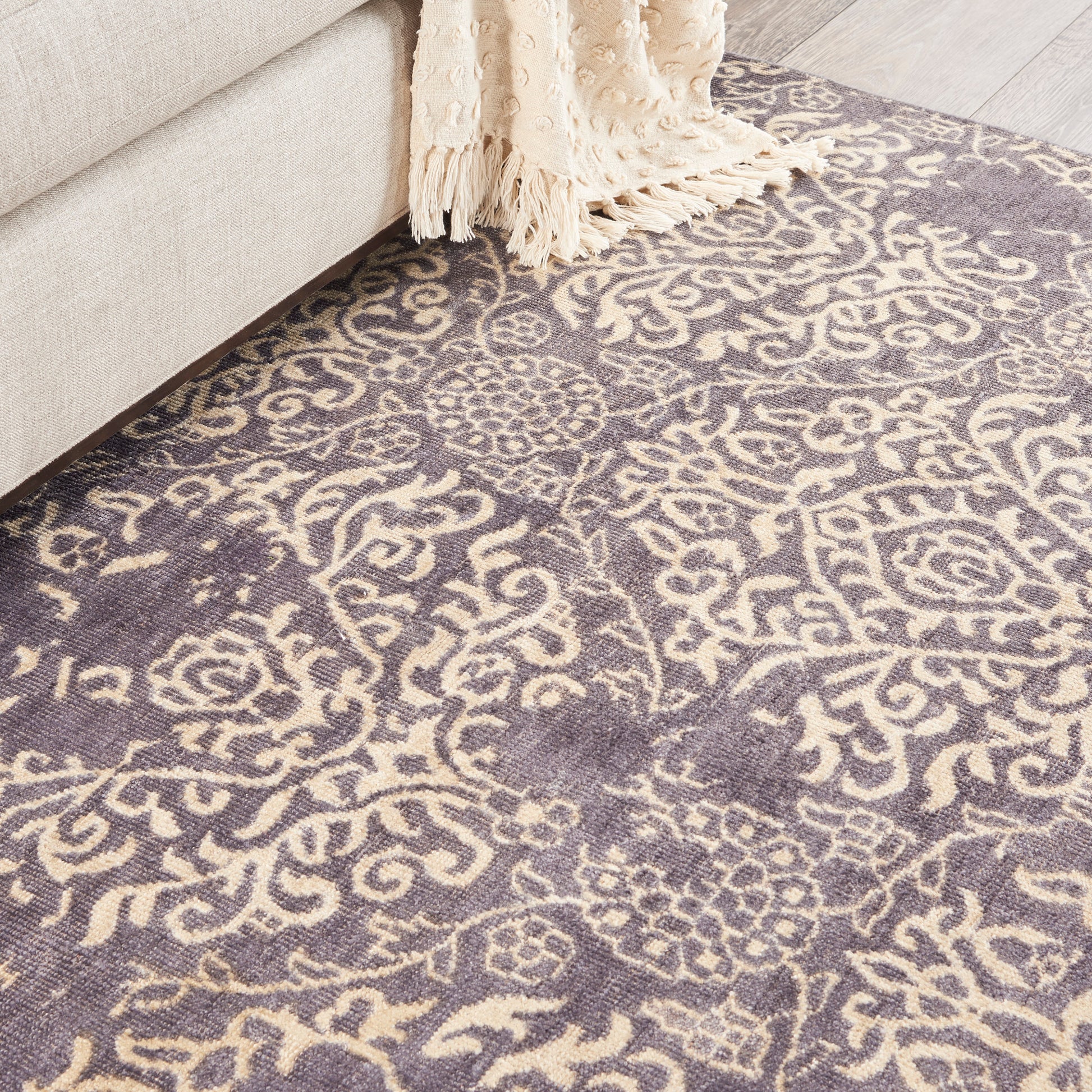 Purple and cream damask pattern rug beside a beige sofa with a cream fringed throw.