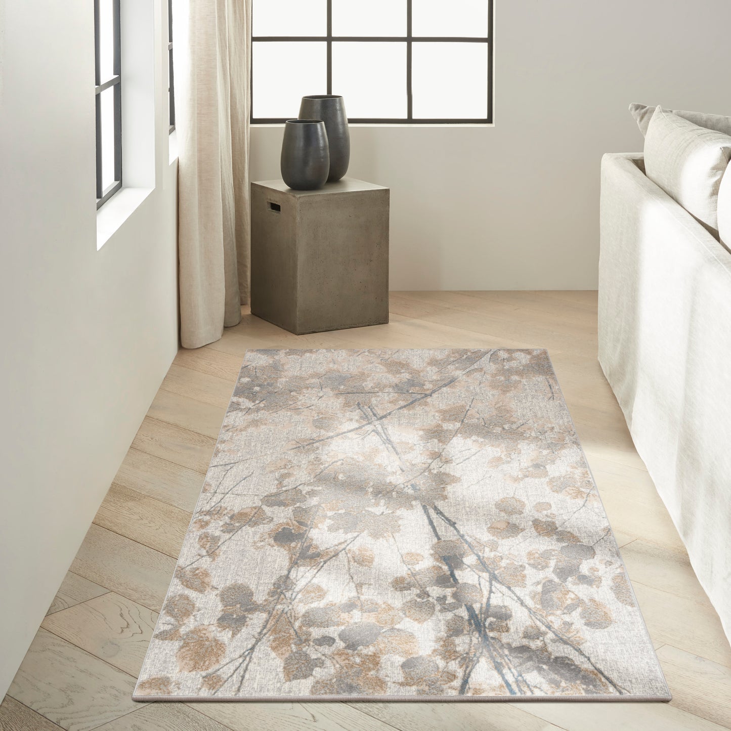 Rectangular neutral area rug with soft beige-taupe-gray botanical pattern on light wood floor in a minimalist living room.