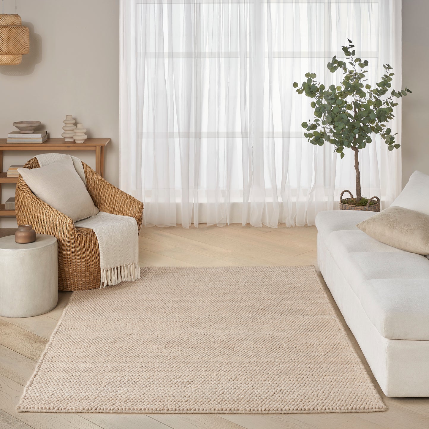 Beige rectangular woven rug in a bright neutral living room with white sofa, rattan armchair, potted tree and sheer curtains.
