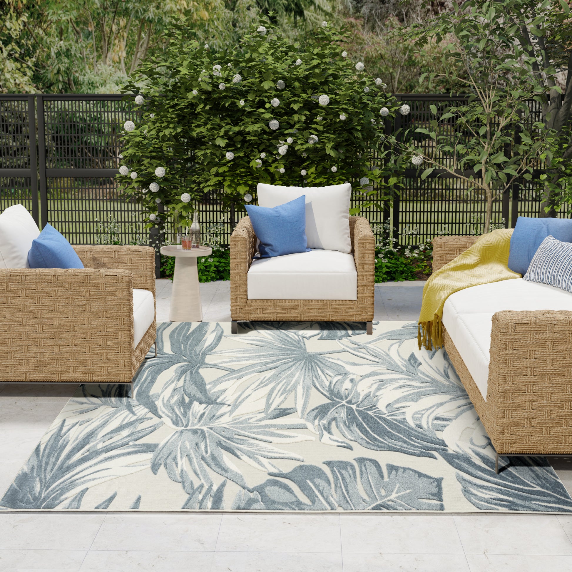Outdoor wicker sofa set with cream cushions, blue pillows, and yellow throw on a palm-leaf patterned rug.