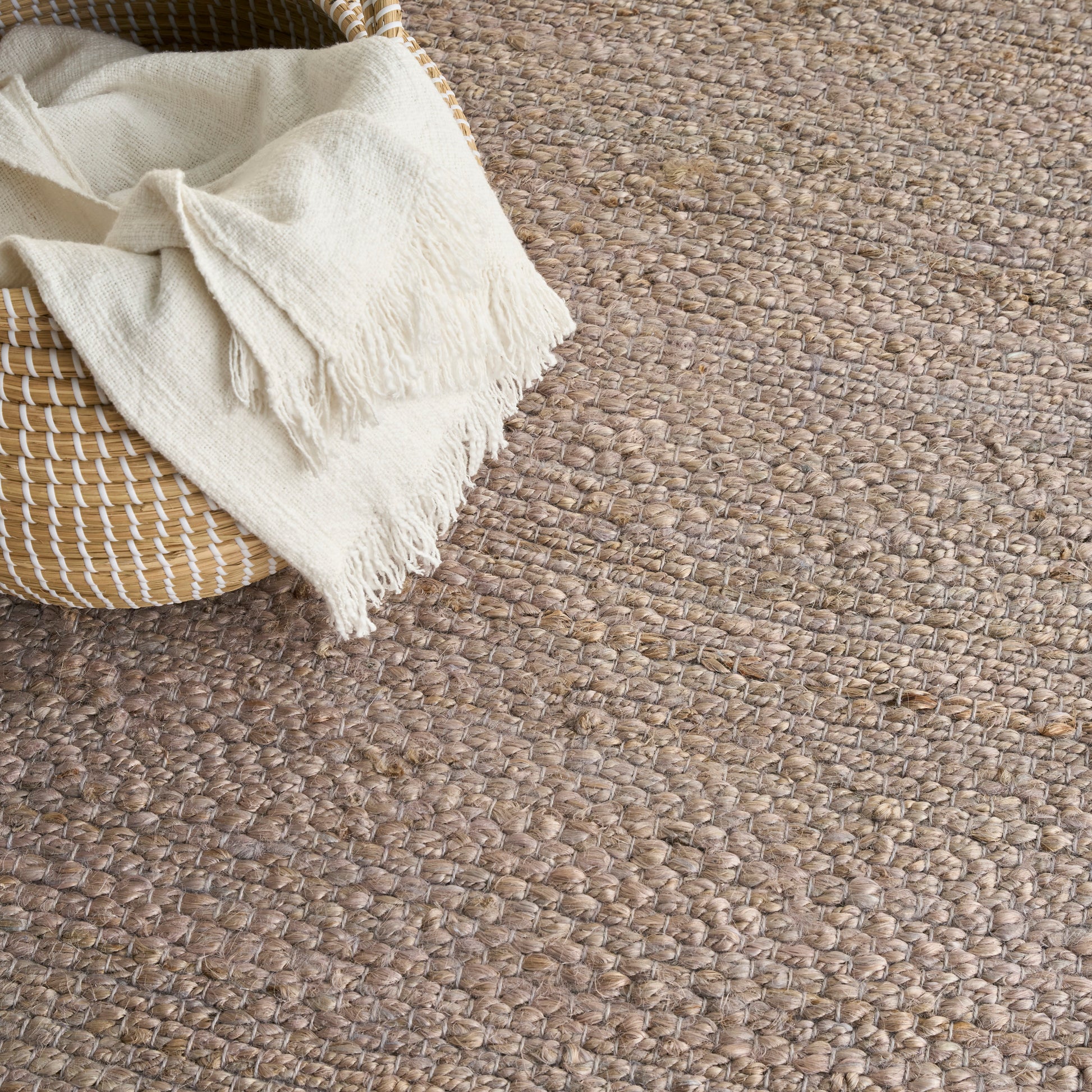 Natural beige jute rug with chunky woven texture; cream fringed throw draped over a round woven basket.