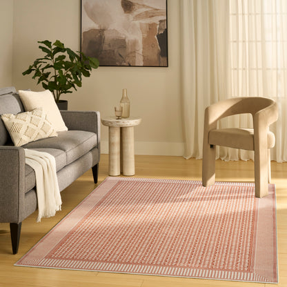 Soft pink geometric patterned area rug in a cozy modern living room with gray sofa, beige chair, plant, and abstract art.