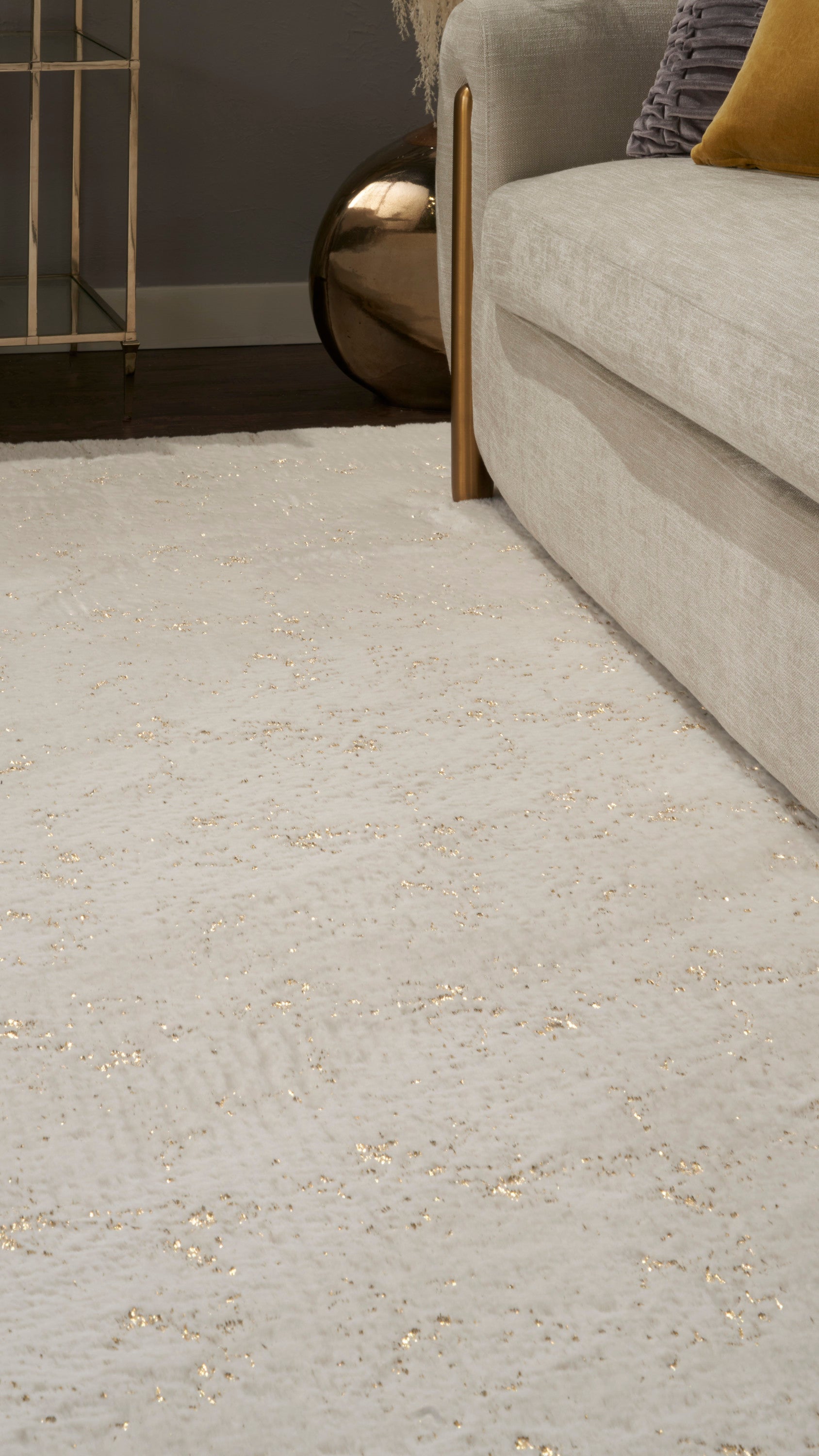 Cream textured rug with subtle gold flecks beside a light taupe sofa and wooden legs; mustard pillow and brass side table.
