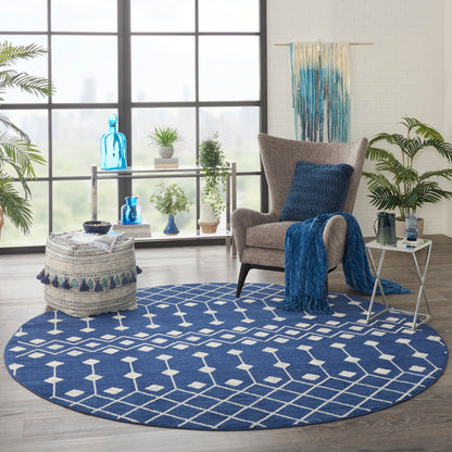Round blue geometric rug anchors a cozy living room with gray armchair, blue throw, cushion, woven pouf, side table and potted plants.