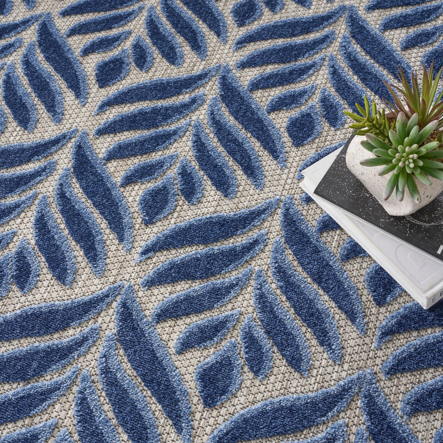 Round beige woven rug with bold blue leaf motifs; tufted texture, modern botanical style, shown with a succulent.