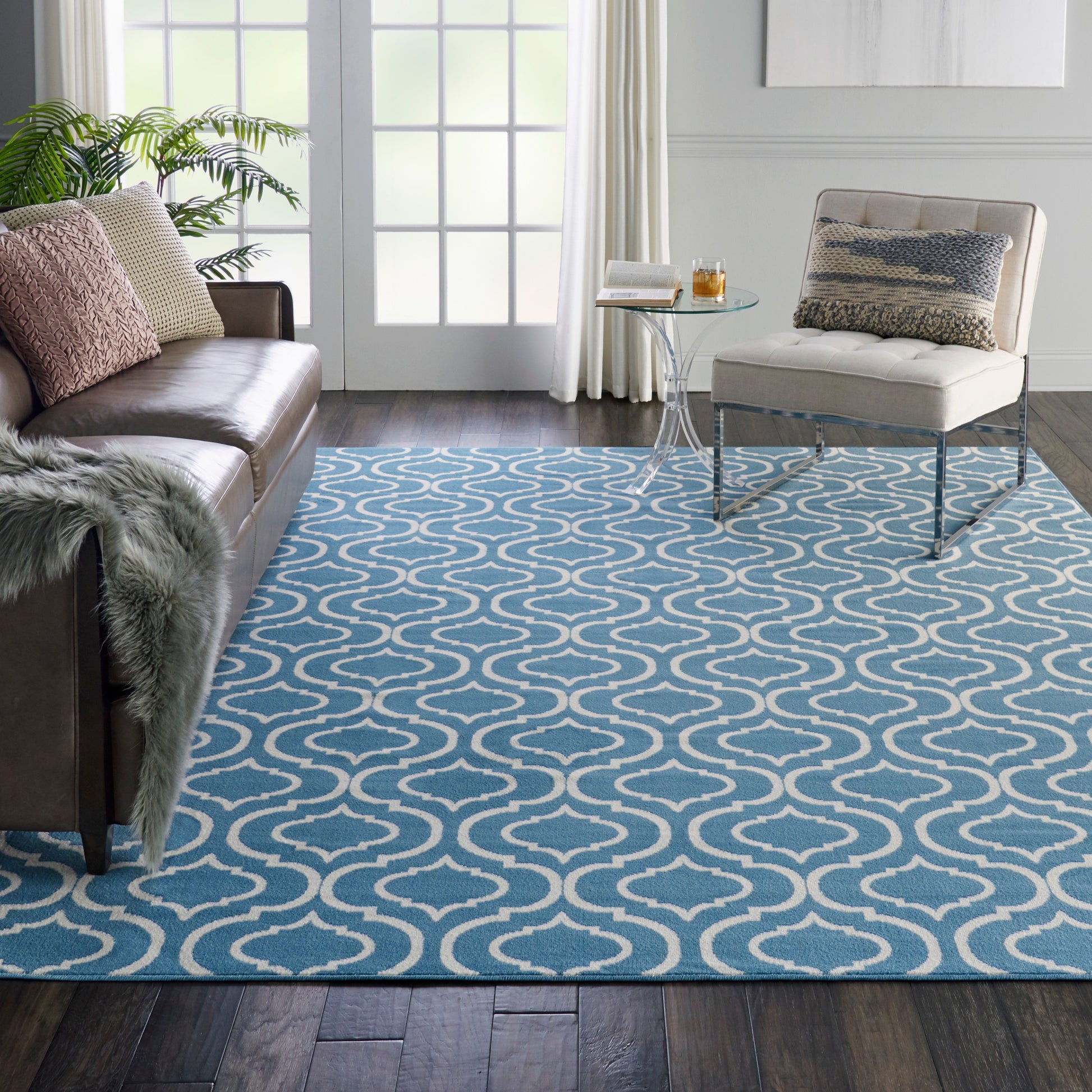 Blue and white arabesque rug in a bright living room with a brown leather sofa, cream chair, fur throw, and glass side table.