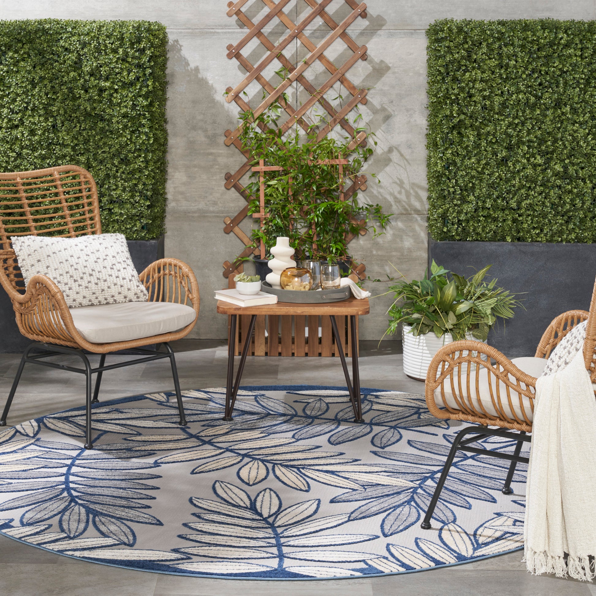 Outdoor wicker lounge set with beige cushions, round blue leaf-pattern rug, wooden coffee table, trellis and hedges
