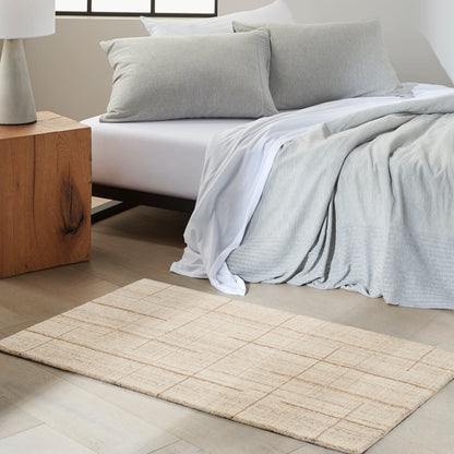 Neutral modern bedroom with white bedding, light gray duvet and pillows, beige woven rectangular rug, and a wooden block nightstand.