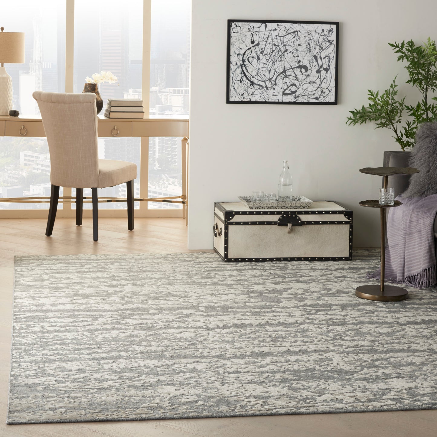 Modern living room with gray abstract rug, beige upholstered chair, studded trunk coffee table, plant, and black-and-white wall art.