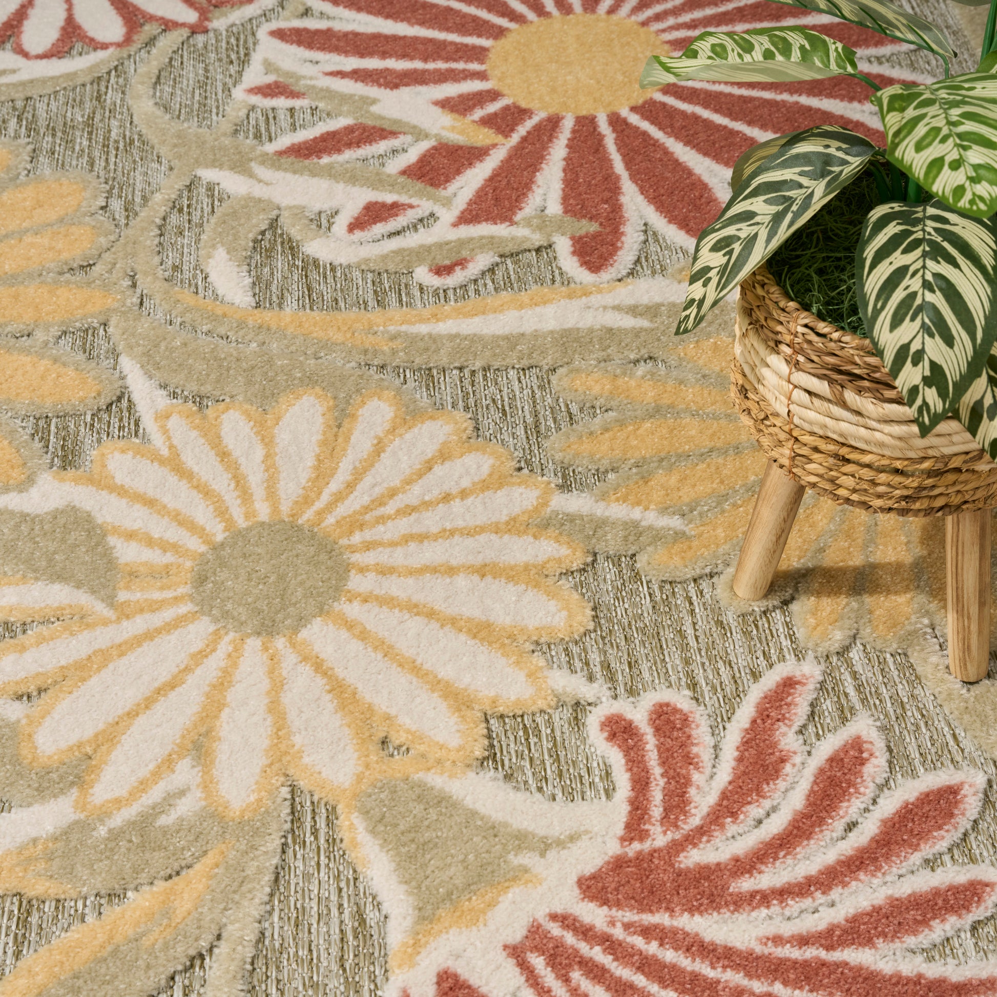 Floral outdoor rug featuring oversized cream-yellow daisies and red petals on a muted gray-green background; woven texture.