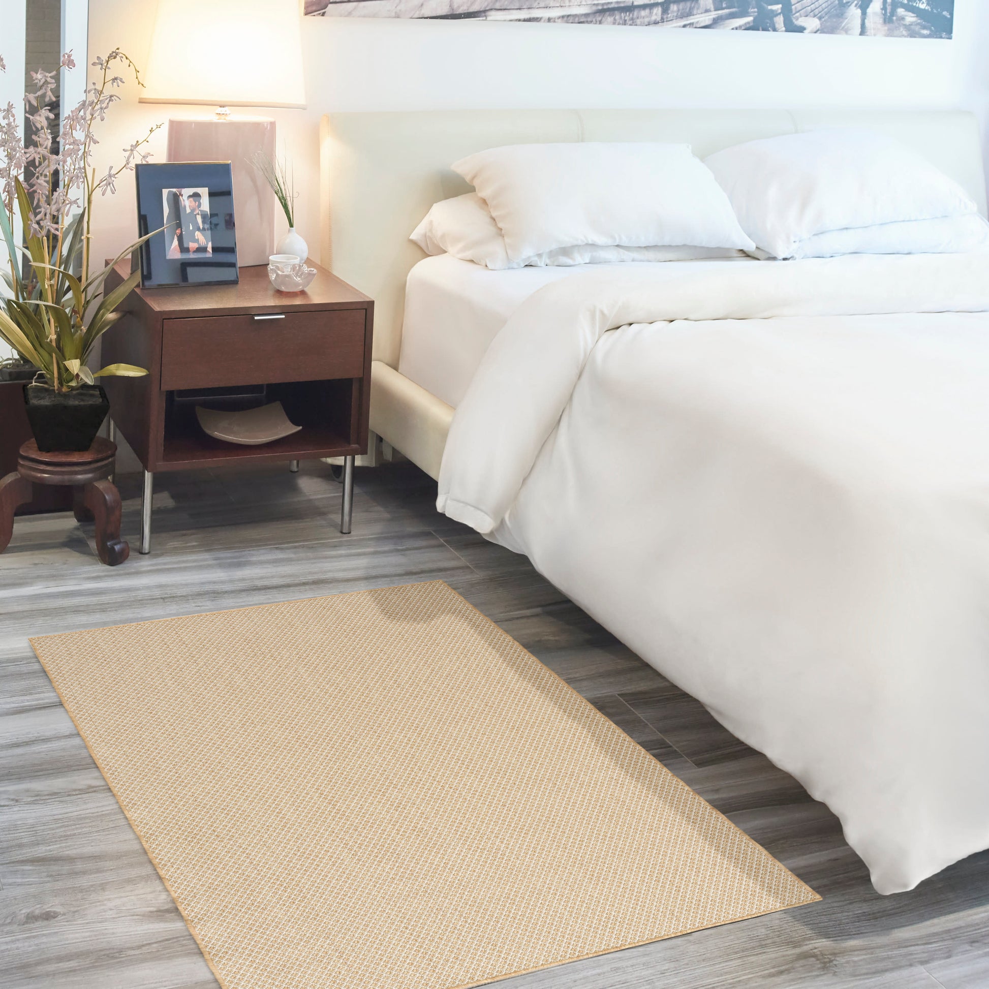 Beige woven rug on gray wood floor beside a white bed with pillows; dark wood nightstand with a lamp, plant, and photo frame.