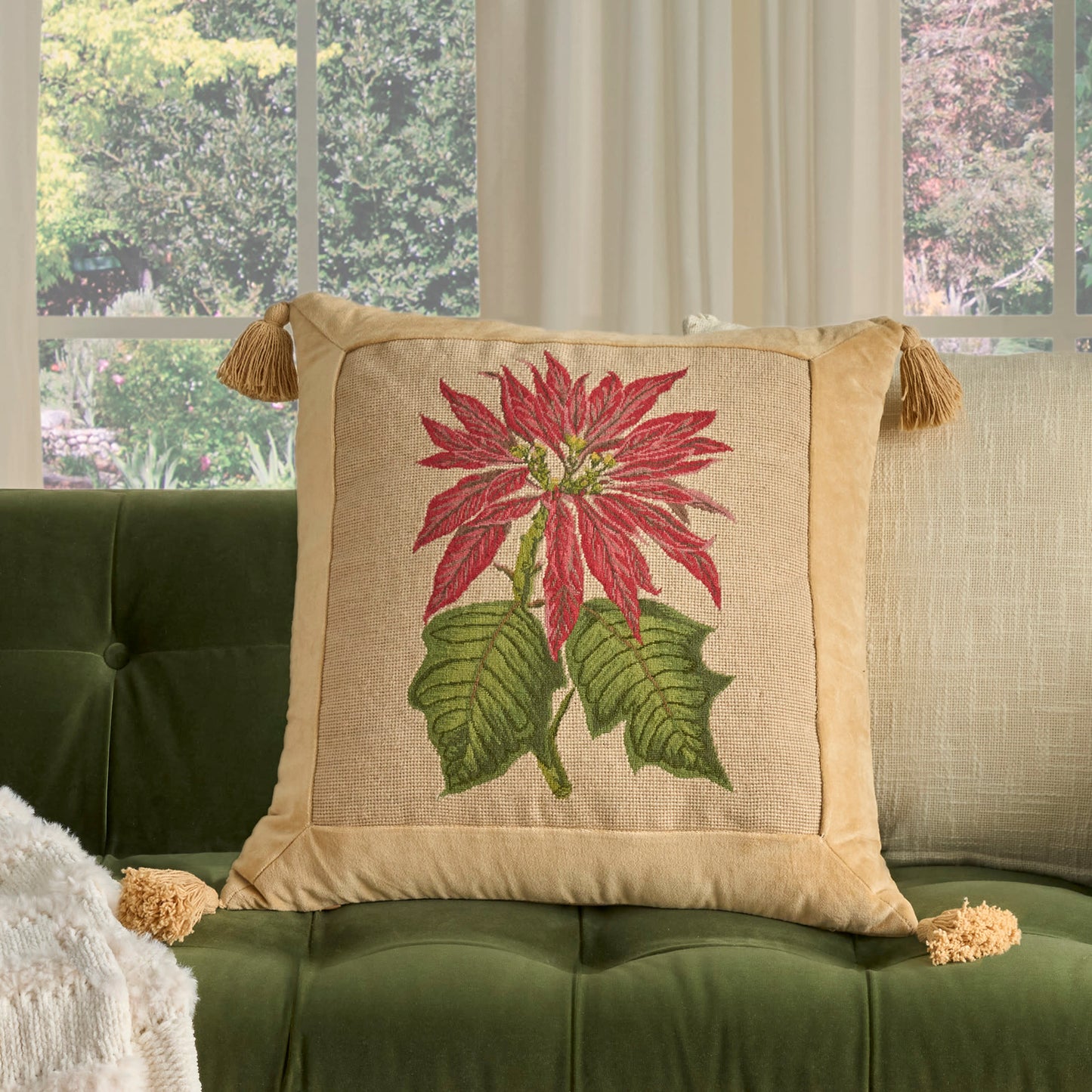 Beige linen decorative pillow with red floral embroidery and tassel-edged corners on a green tufted sofa.