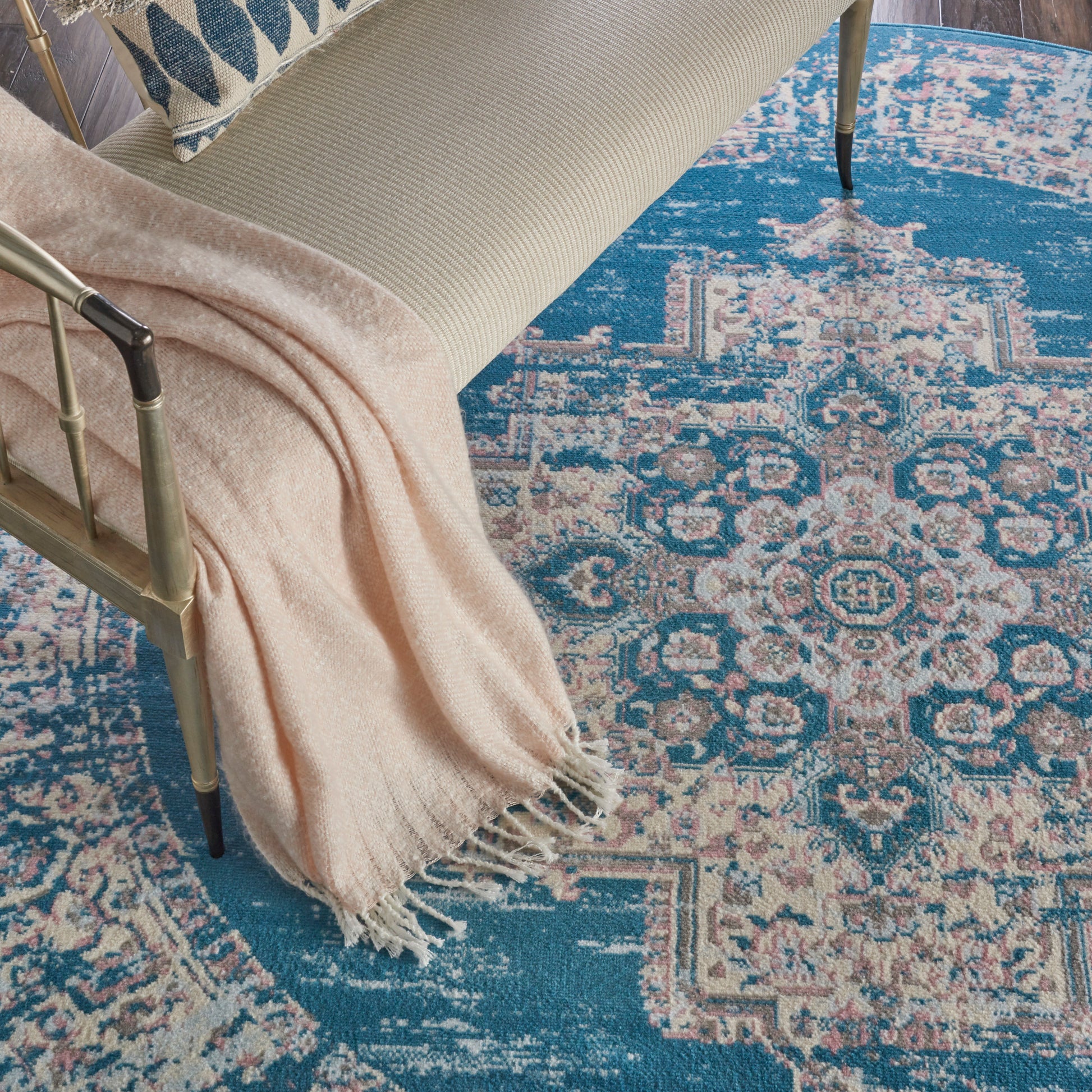Beige upholstered chair with a blush pink fringed throw, gold frame, atop a teal distressed oriental rug.