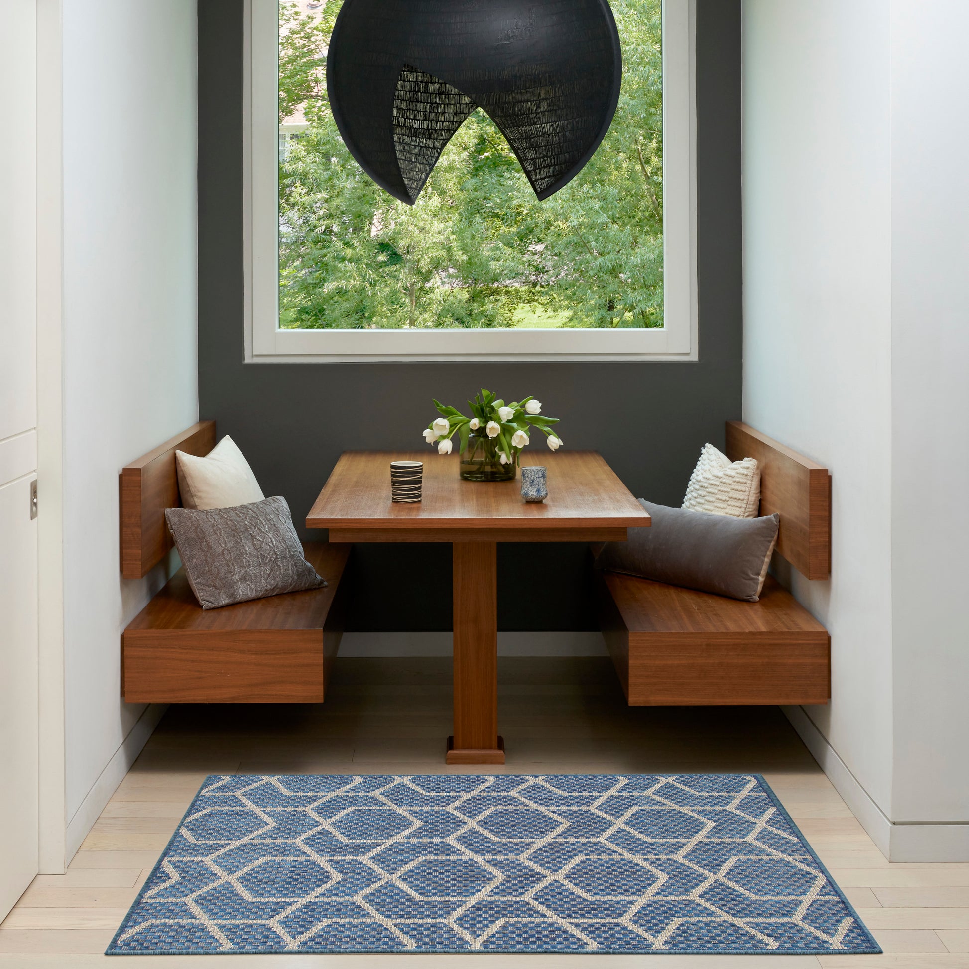 Wooden corner dining nook with square table, twin benches, gray wall, blue geometric rug, black pendant lamp, white flowers.
