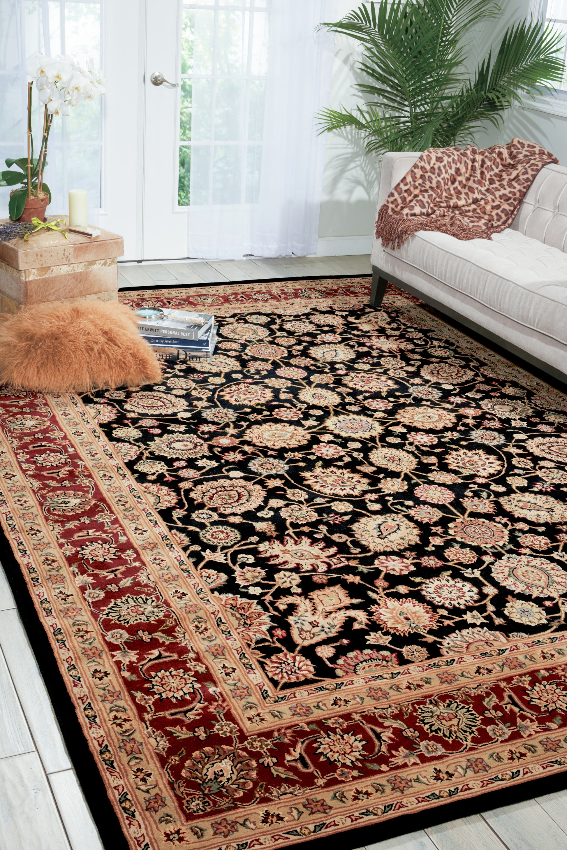Large rectangular Persian-style rug with a black field and red and beige floral border; white sofa with a leopard-print throw, and plants nearby.