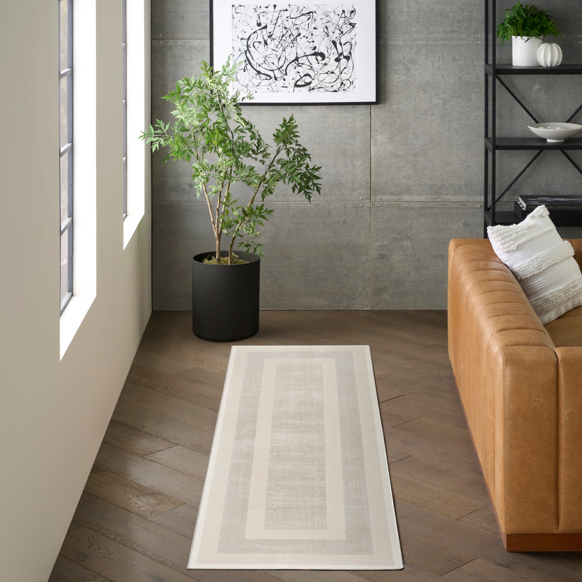 Light-gray runner rug with double border beside a tan leather sofa, green potted plant & black metal shelf.
