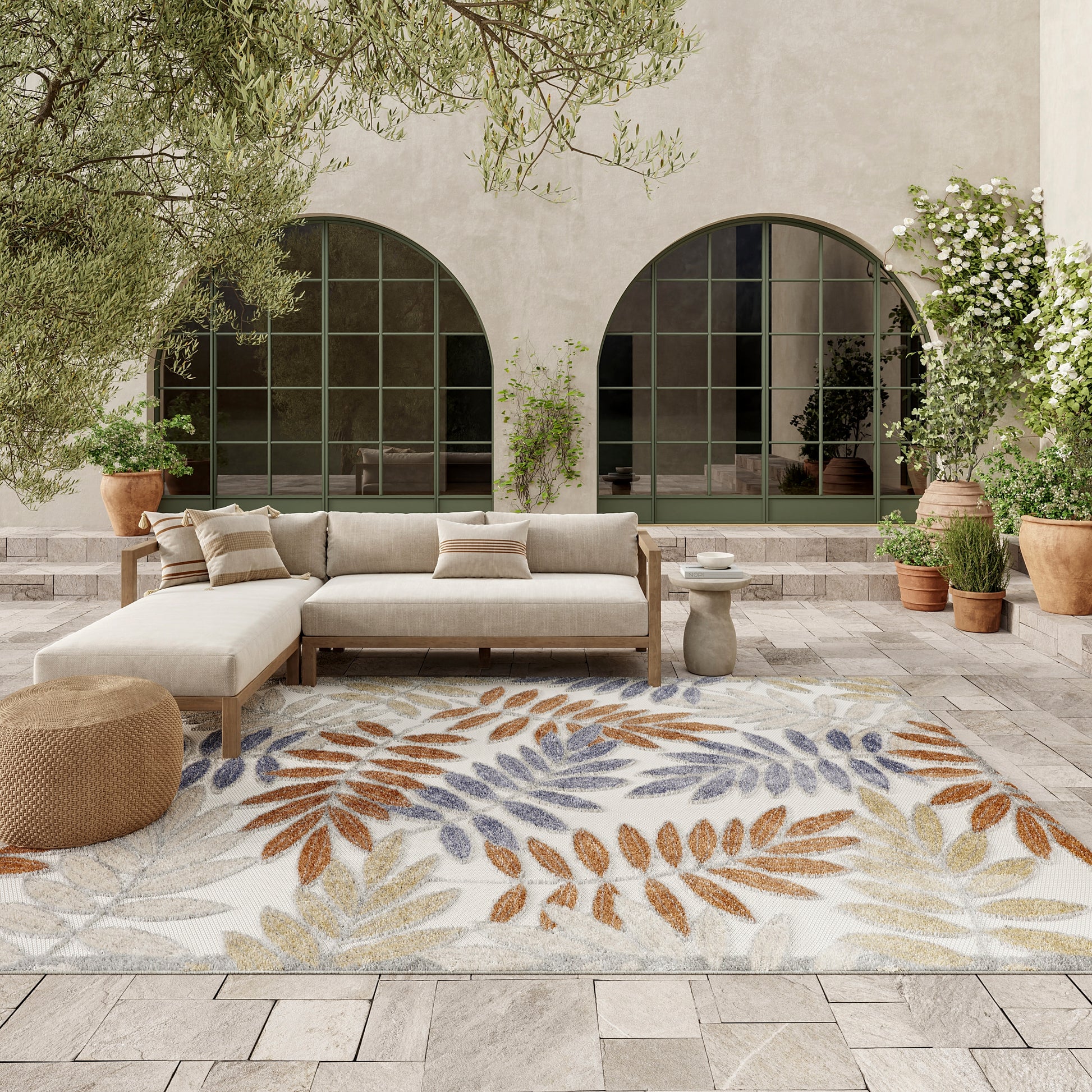 Beige outdoor sectional on leaf-pattern rug with orange, blue, and yellow leaves, arched windows, and potted plants.