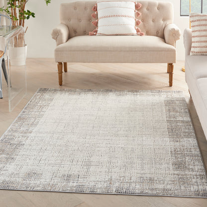 Beige tufted loveseat with wooden legs, pink-fringe pillows, and a gray distressed-area rug in a bright living room.