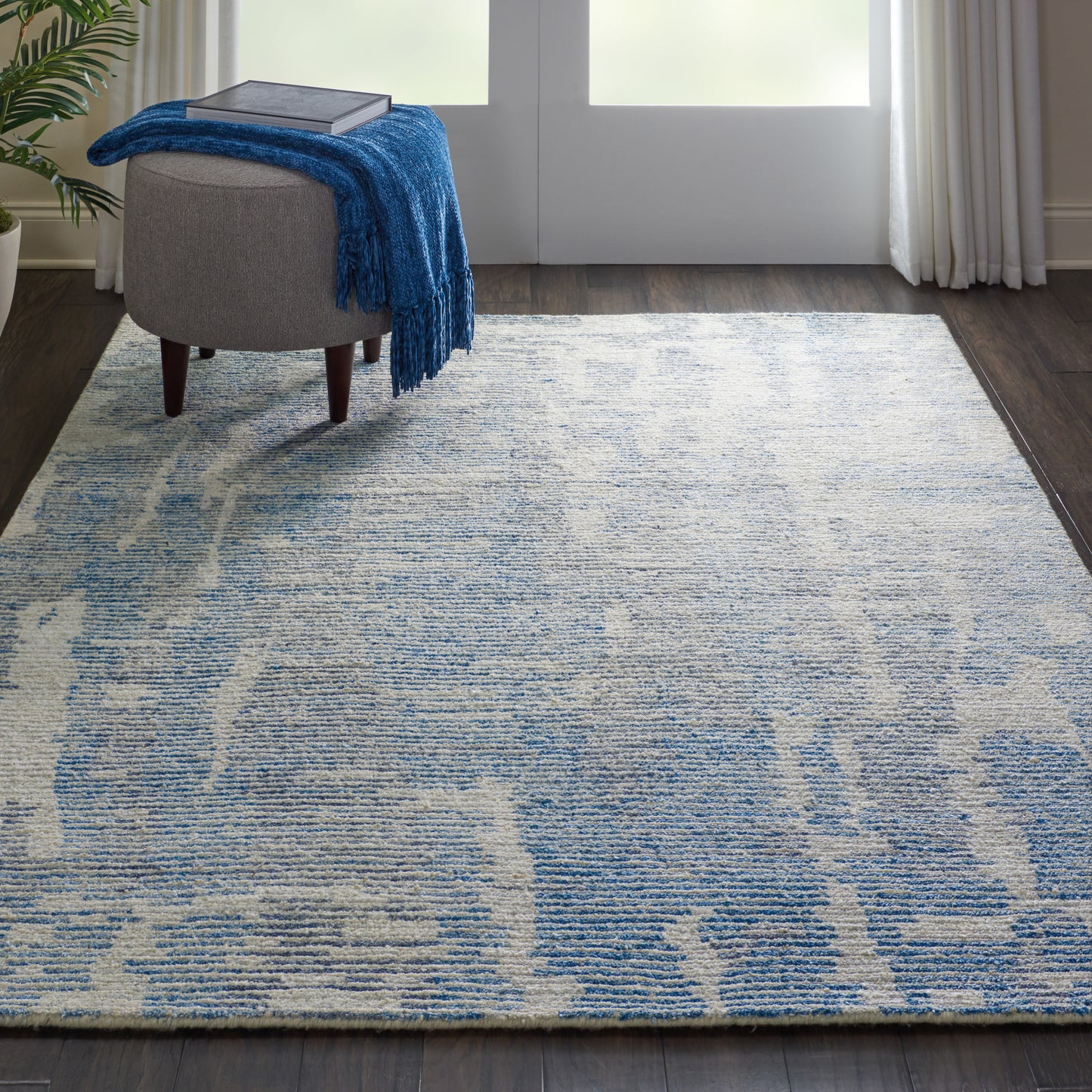 Blue and ivory abstract-pattern area rug on dark hardwood, with a gray ottoman and blue throw blanket.