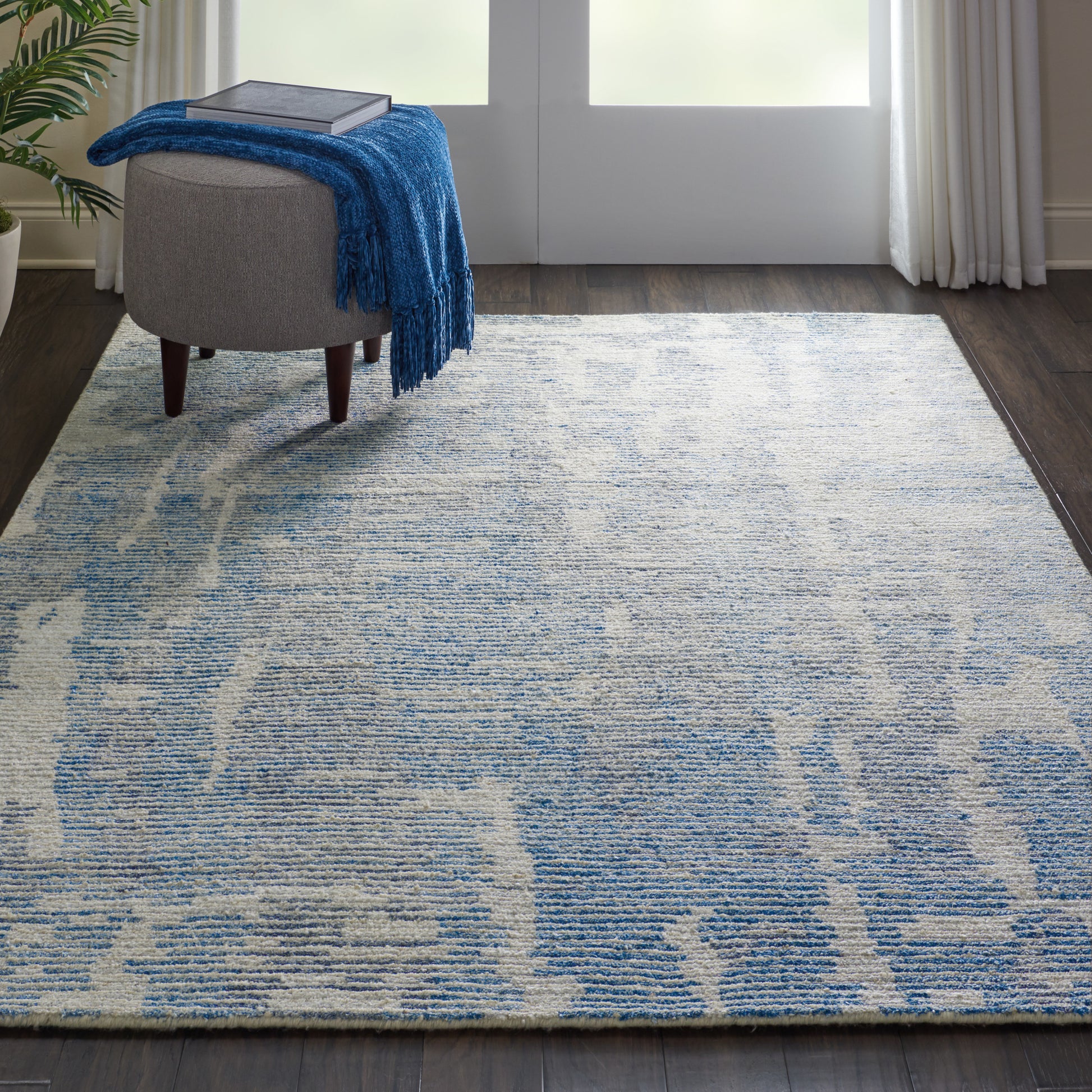 Blue and ivory abstract-pattern area rug on dark hardwood, with a gray ottoman and blue throw blanket.