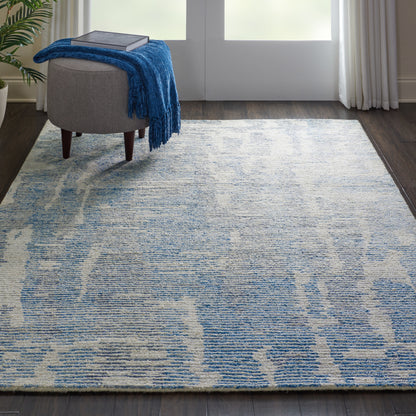 Blue and ivory abstract-pattern area rug on dark hardwood, with a gray ottoman and blue throw blanket.