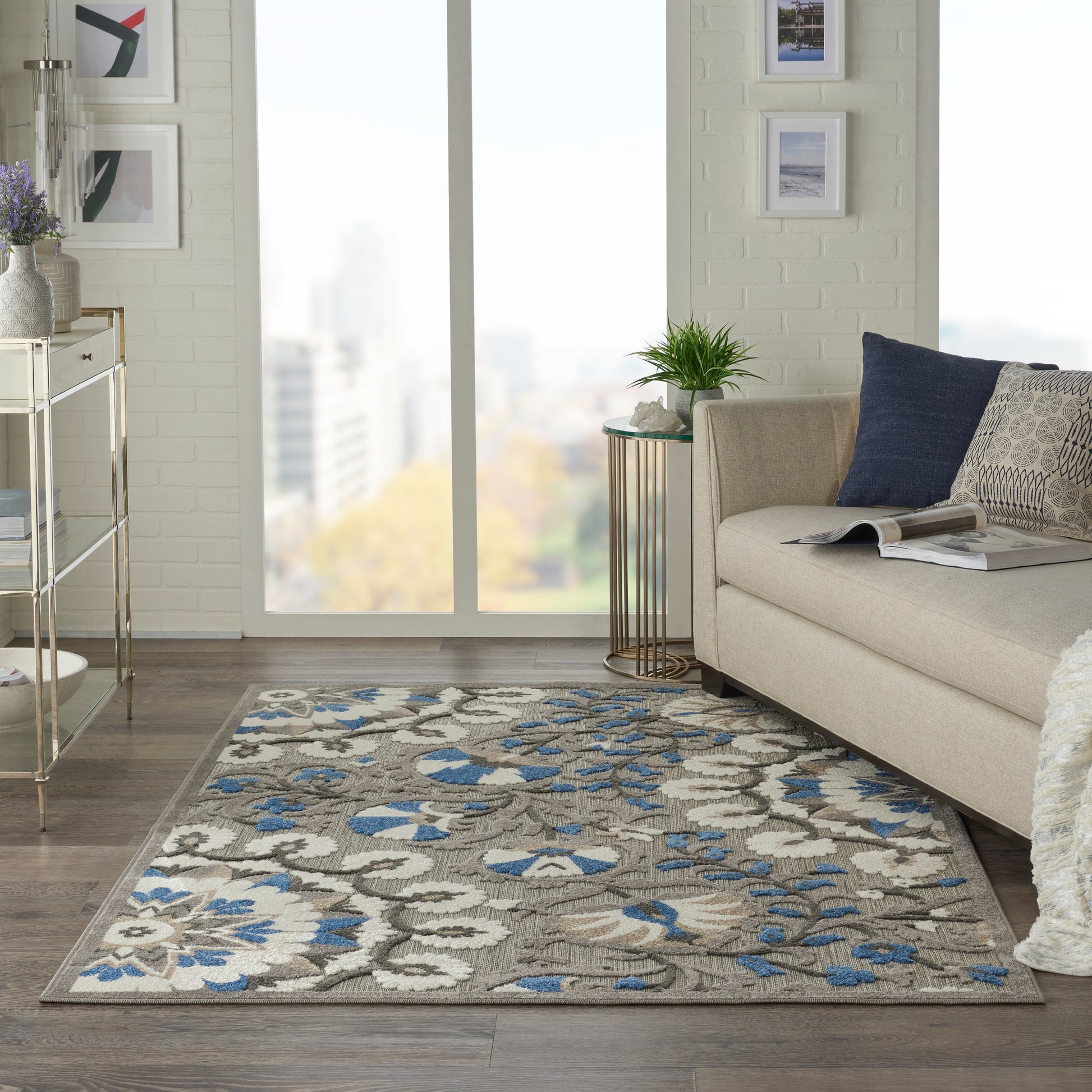 Rectangular beige-gray area rug with blue floral motif, set in a modern living room beside a beige sofa and large window.
