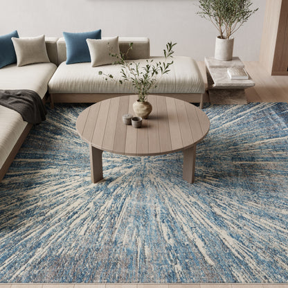 Blue abstract rug on the floor with a round wooden coffee table, beige sectional sofa & blue cushions, and a potted plant.