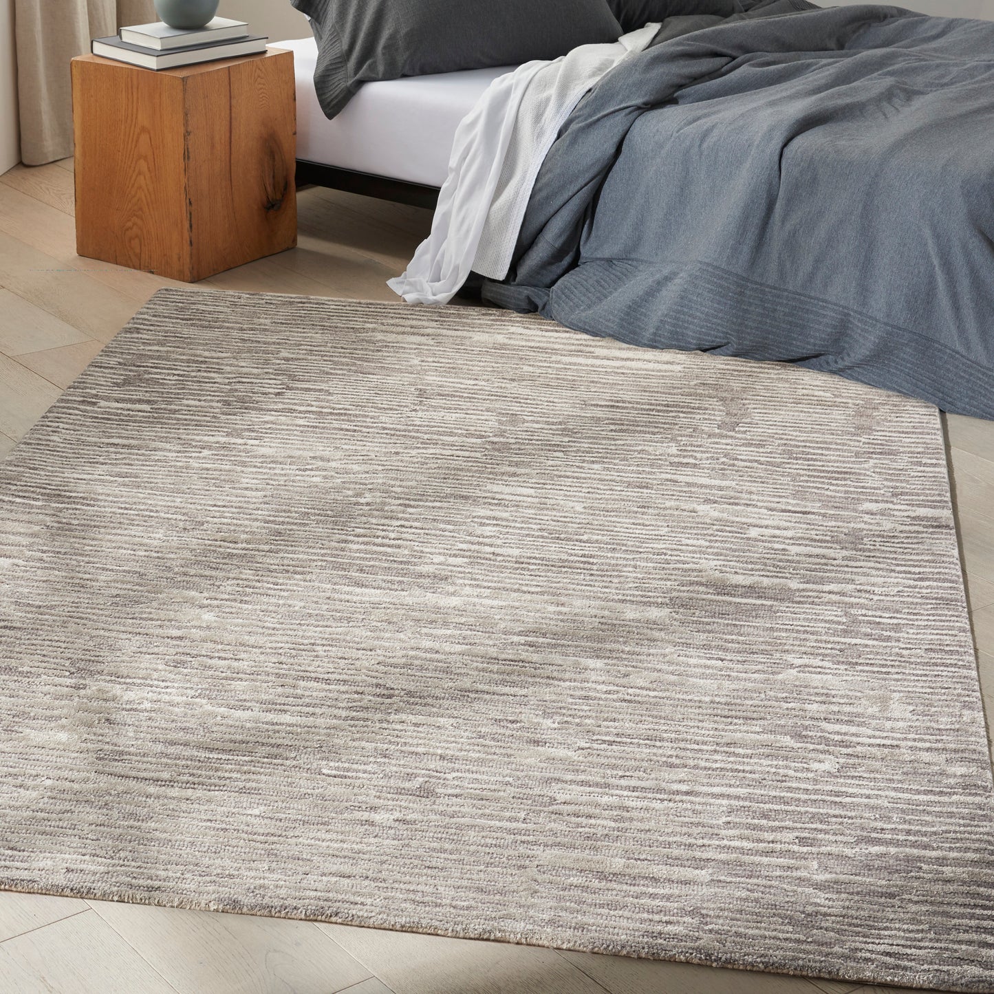 Neutral beige-gray textured area rug with subtle lines in a bedroom beside a wooden nightstand and gray bedding.